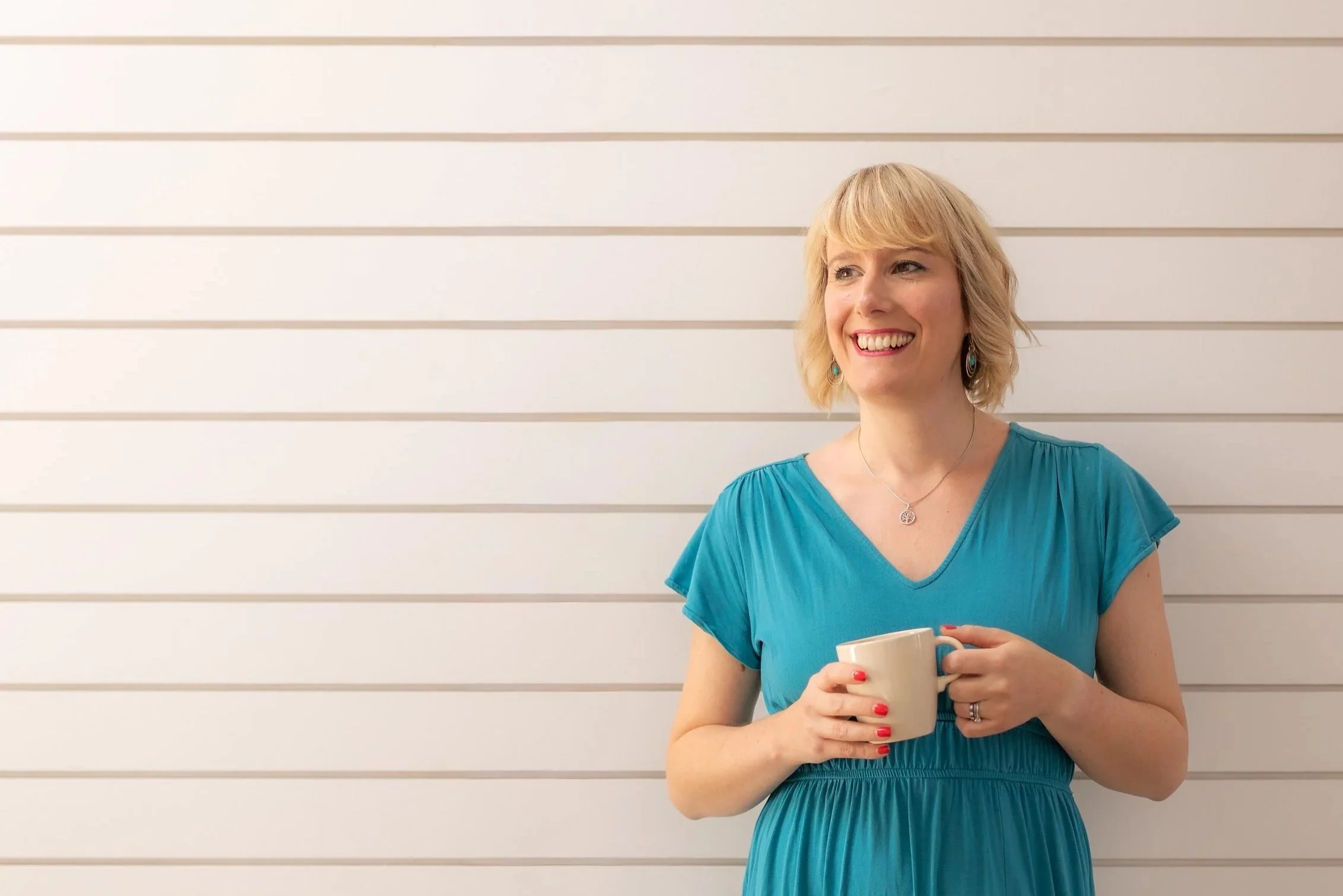 A woman with blonde hair and a big smile, wearing a blue dress, holding a coffee mug, standing against a white horizontal-striped wall.