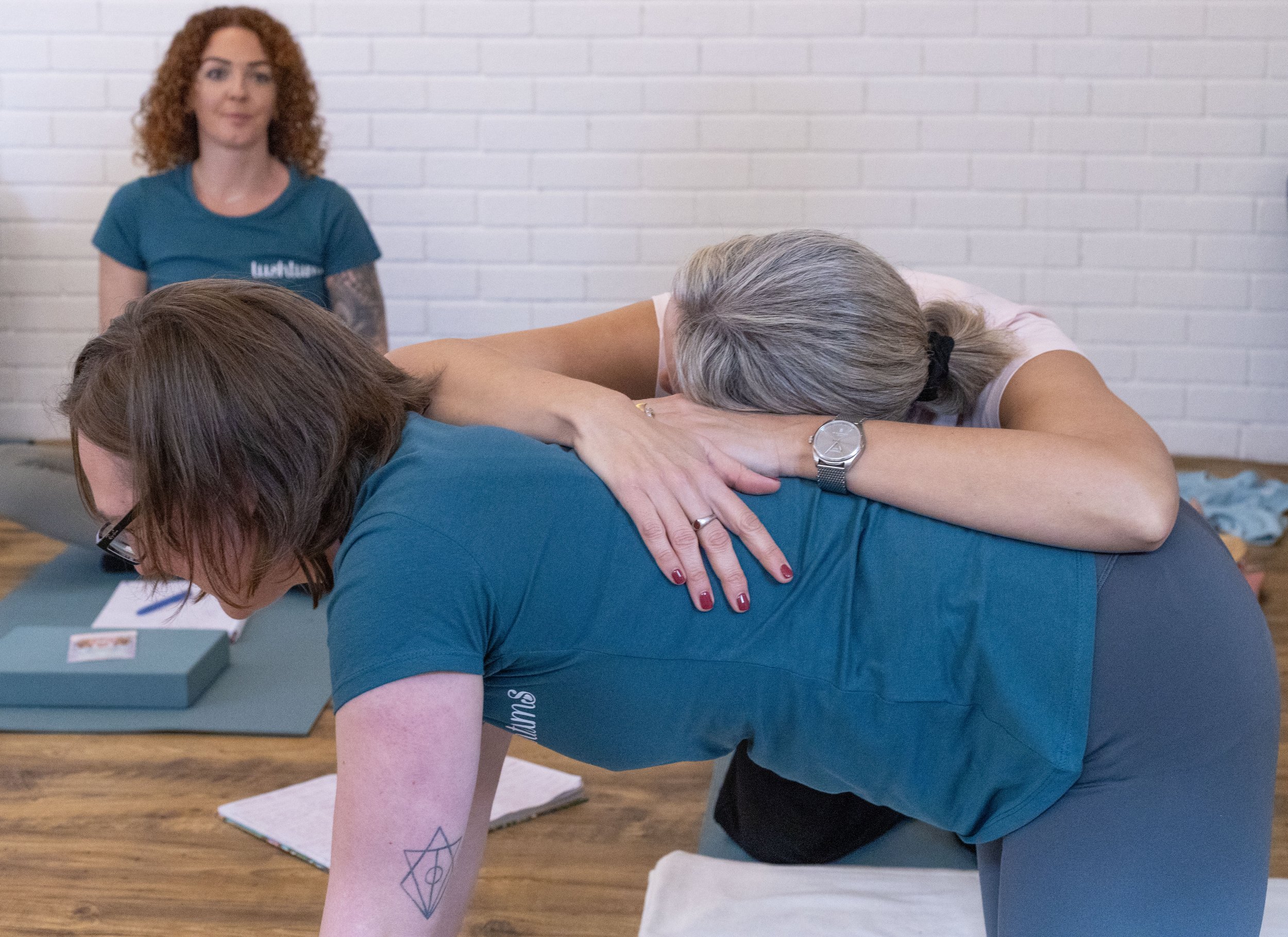 Two women practicing yoga or massage, one woman leaning over a table while the other supports her from behind, with a third woman in the background observing.