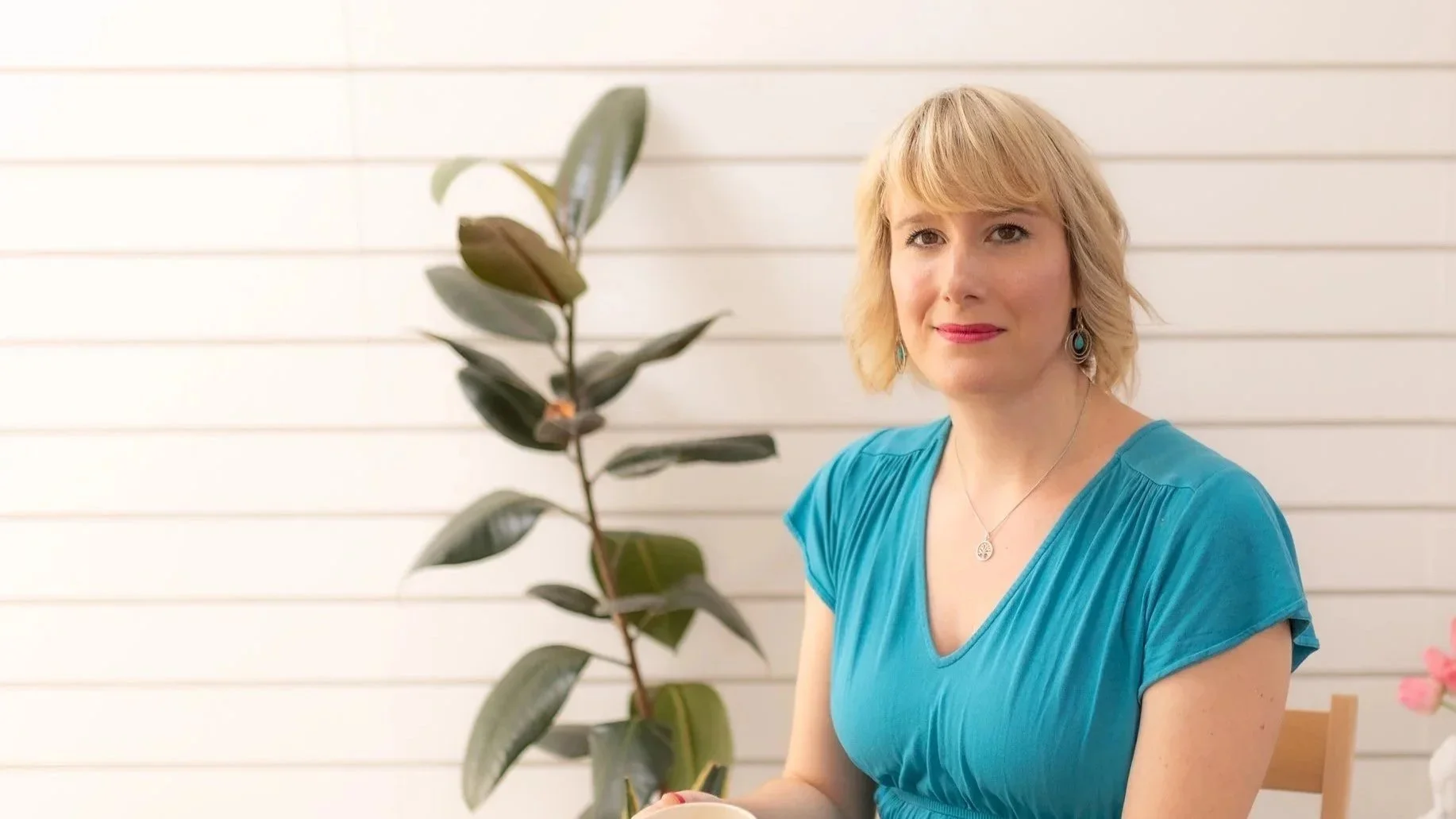 A woman with blonde hair wearing a turquoise shirt and turquoise earrings, sitting in front of a white background, with a plant in the foreground.
