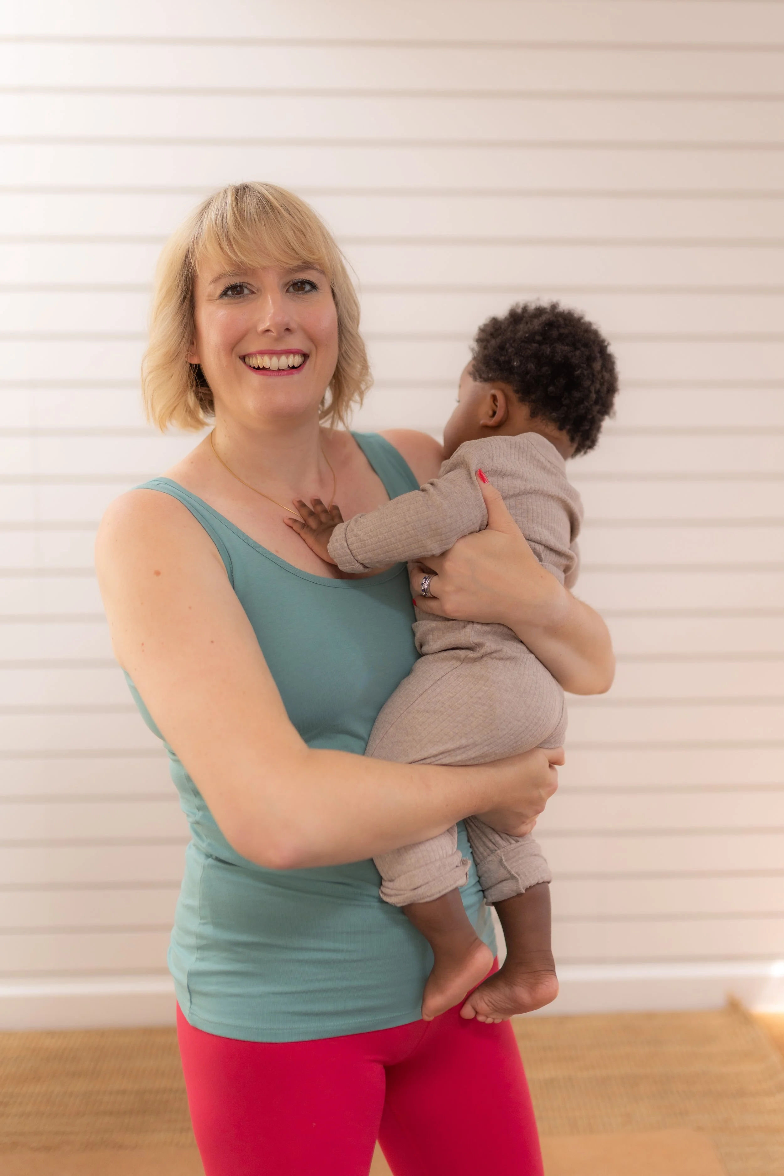 A woman with blonde hair and fair skin holding a young child with dark curly hair and darker skin, both smiling, indoors with white horizontal blinds in the background.