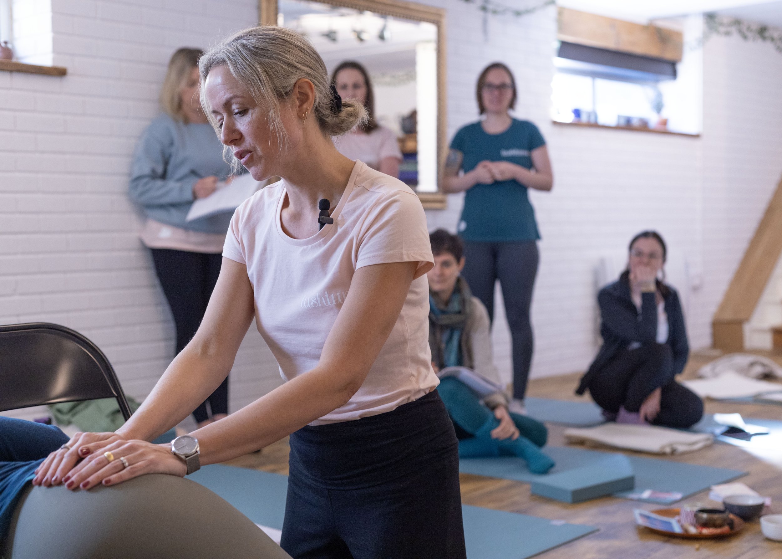 A woman in a white t-shirt demonstrates a yoga pose for pregnancy and birth on a mat in a wellness class, with several women sitting or standing nearby in a brightly lit room with white brick walls.