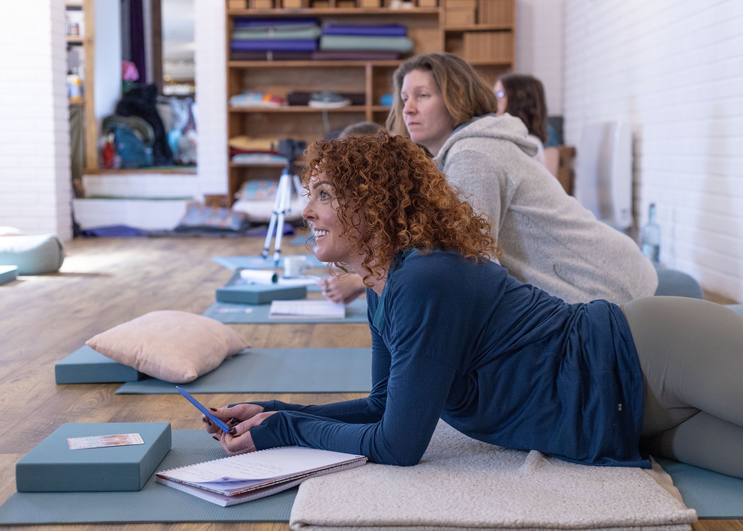 Women participating in a yoga for birth training class, lying on their sides on yoga mats in a studio, with one woman smiling and holding a phone.