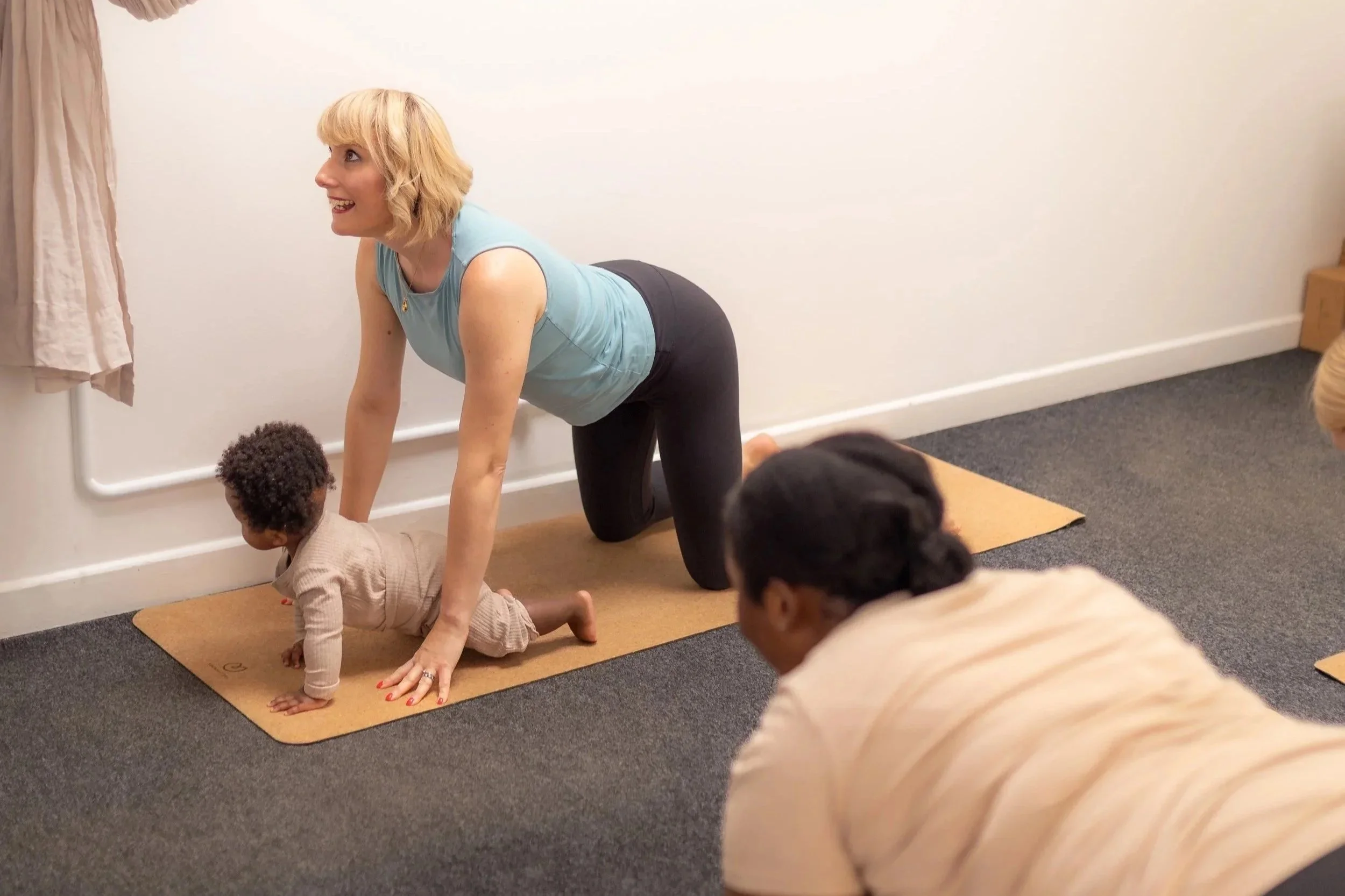 A woman with blonde hair in a teal sleeveless top and black leggings leading a group yoga session on a yoga mat in a room with beige walls. She is on hands and knees in cow pose, with a baby in a similar pose.