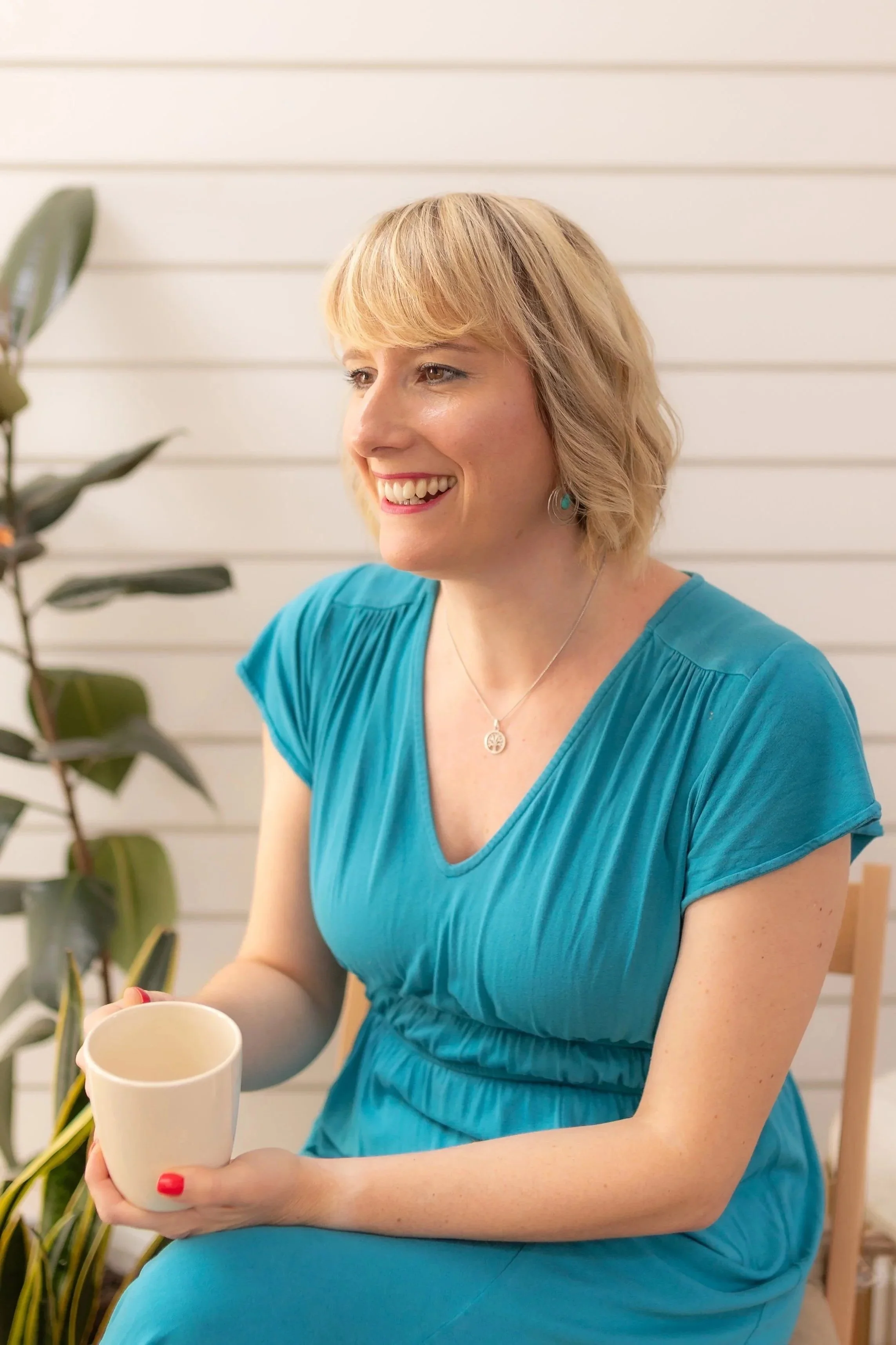 A woman with blonde hair wearing a turquoise dress, sitting at a table, holding a white mug with red nail polish, smiling, in front of a white wall with a large plant nearby.