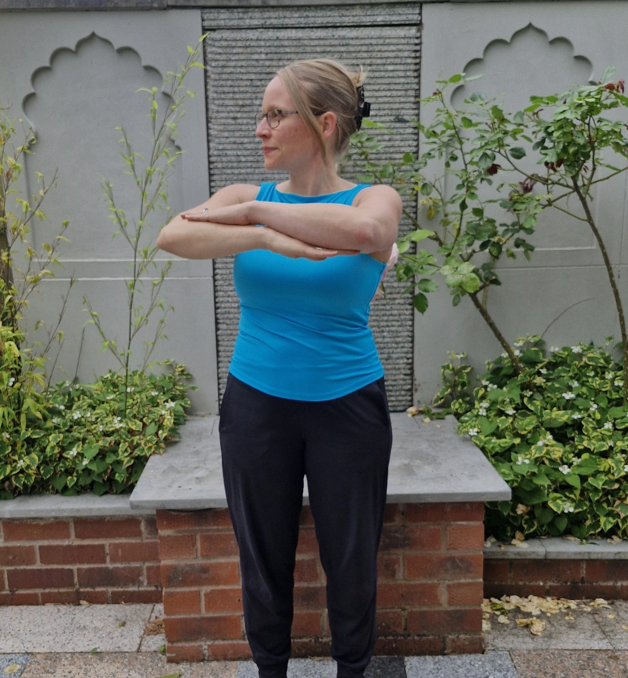 A woman standing outdoors with arms crossed in front of her doing a Pilates waist twist, wearing a blue sleeveless shirt and black pants, with glasses and a black hair clip in her hair.