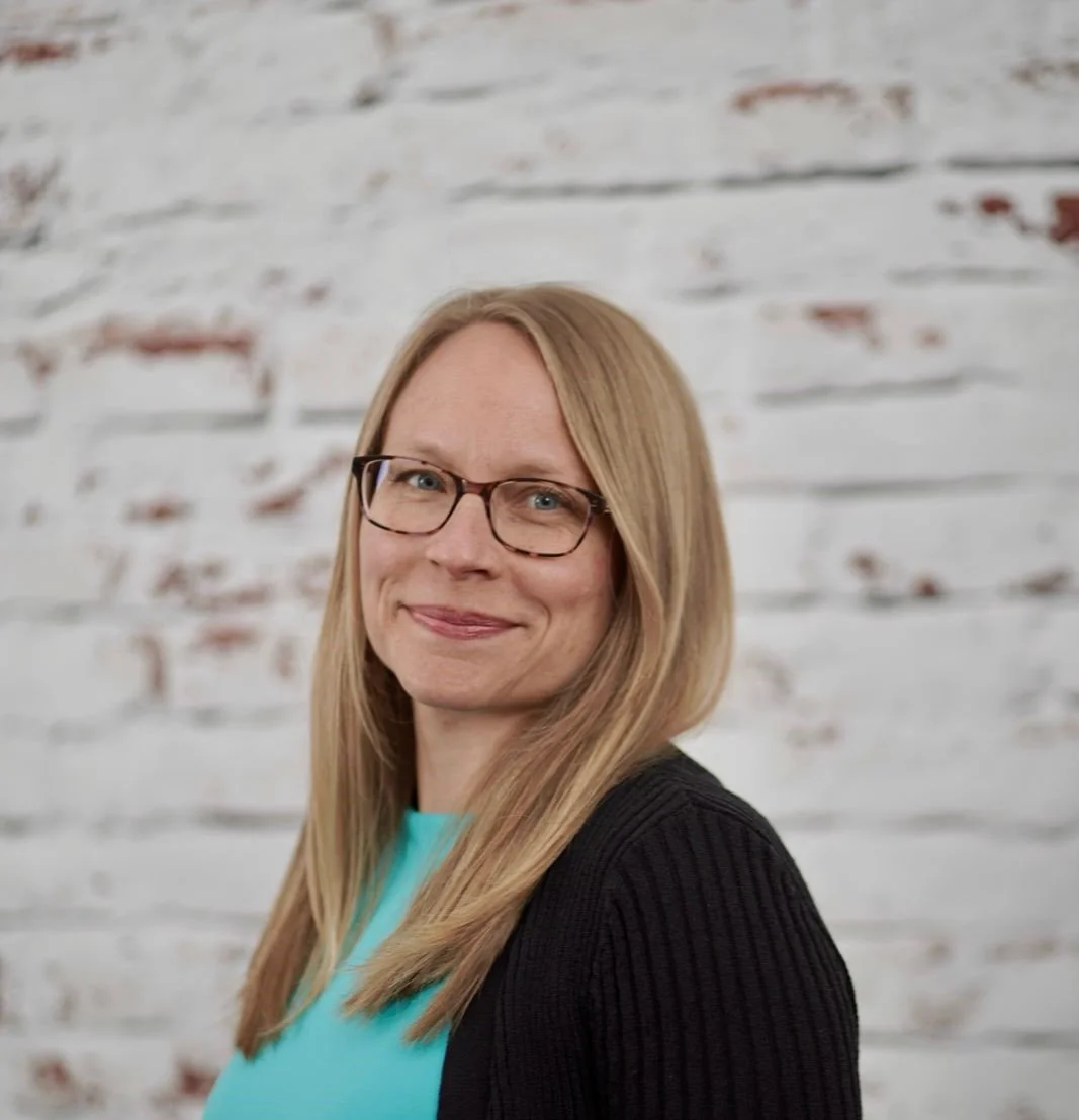 A woman with blonde hair, glasses, and a black sweater, smiling against a white brick wall.