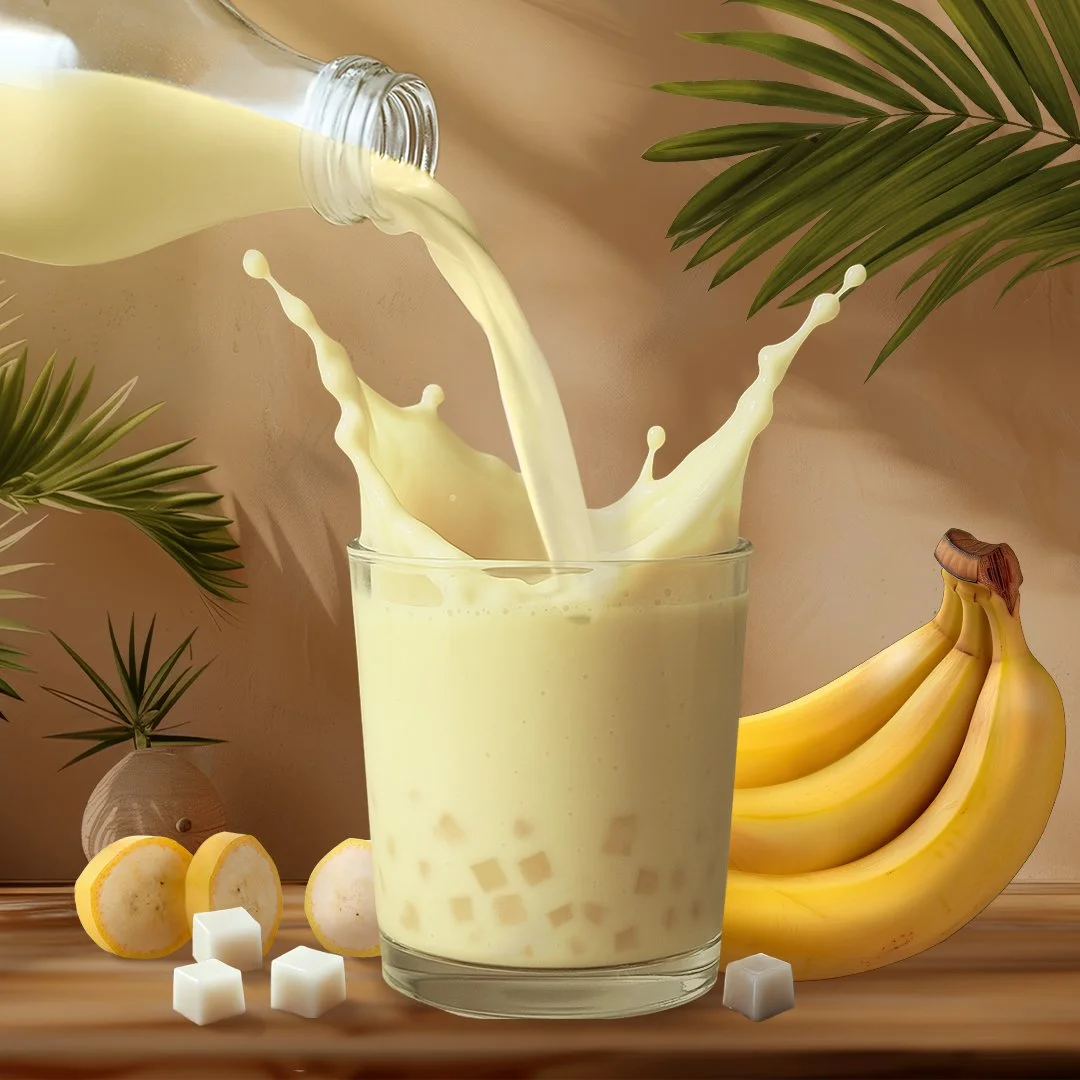 Pouring banana milk into a glass with banana slices, a bunch of bananas, and sugar cubes beside it.