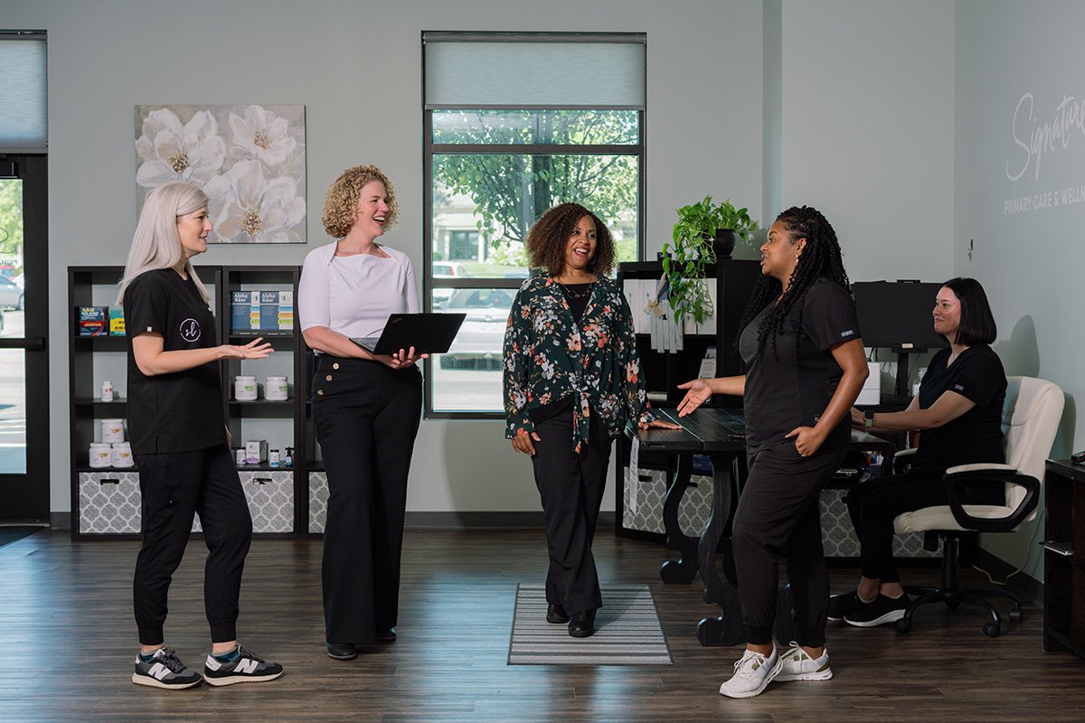 Five women in an office having a conversation, with some standing and others sitting at a desk.