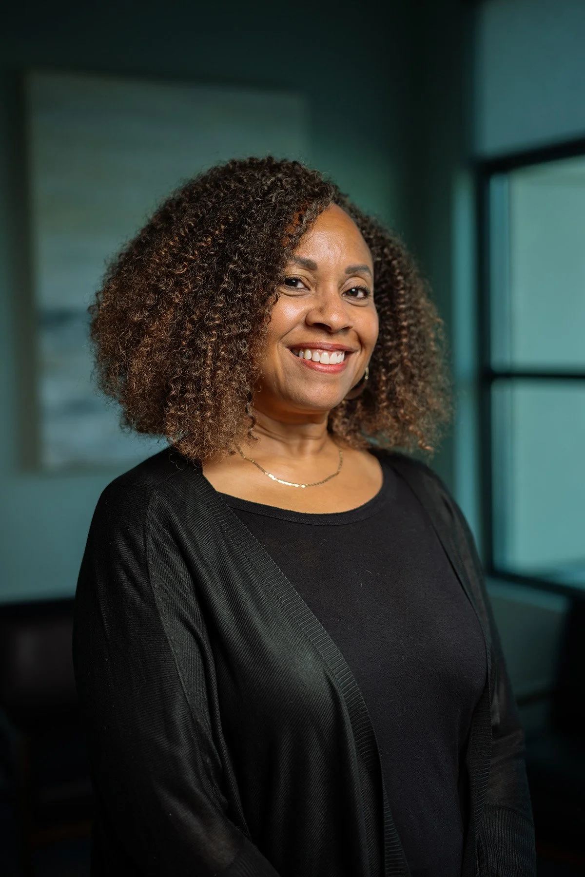 A woman with curly hair smiling, wearing a black top and a delicate gold necklace, indoors near a window.