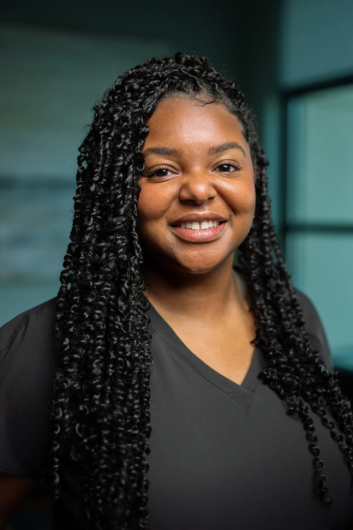 A woman with long, curly black hair smiling in an indoor setting with blurred windows in the background.