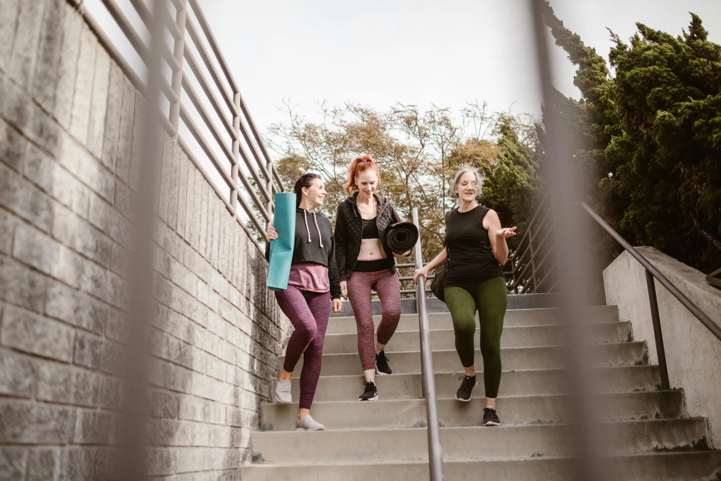 Three women in athletic attire walking down outdoor concrete stairs, carrying yoga mats and talking, with trees and a cloudy sky in the background.