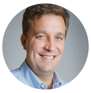 Portrait of a smiling man with short brown hair wearing a light blue collared shirt against a gray background.