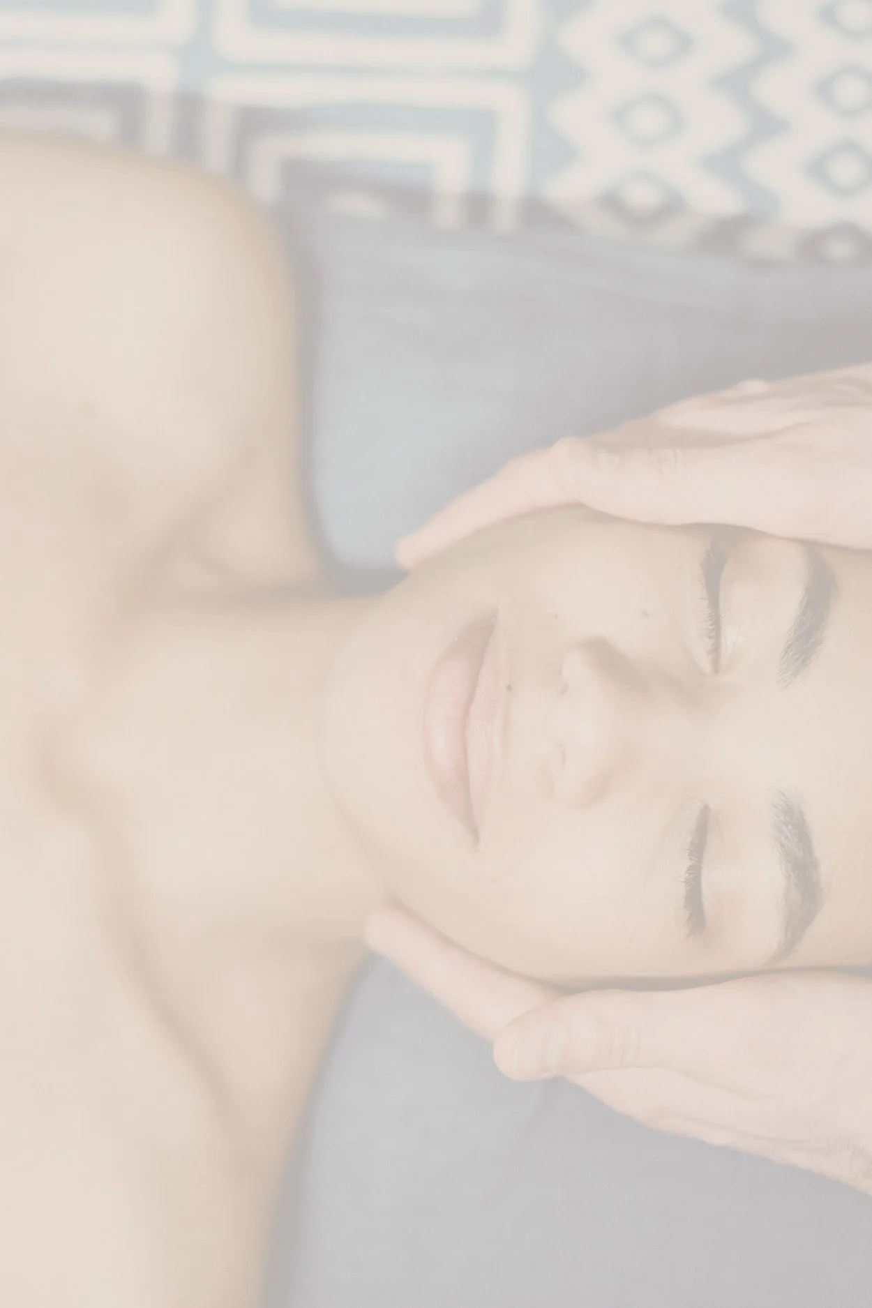 Close-up of a woman with closed eyes receiving a facial massage, lying on a gray surface with a patterned background.