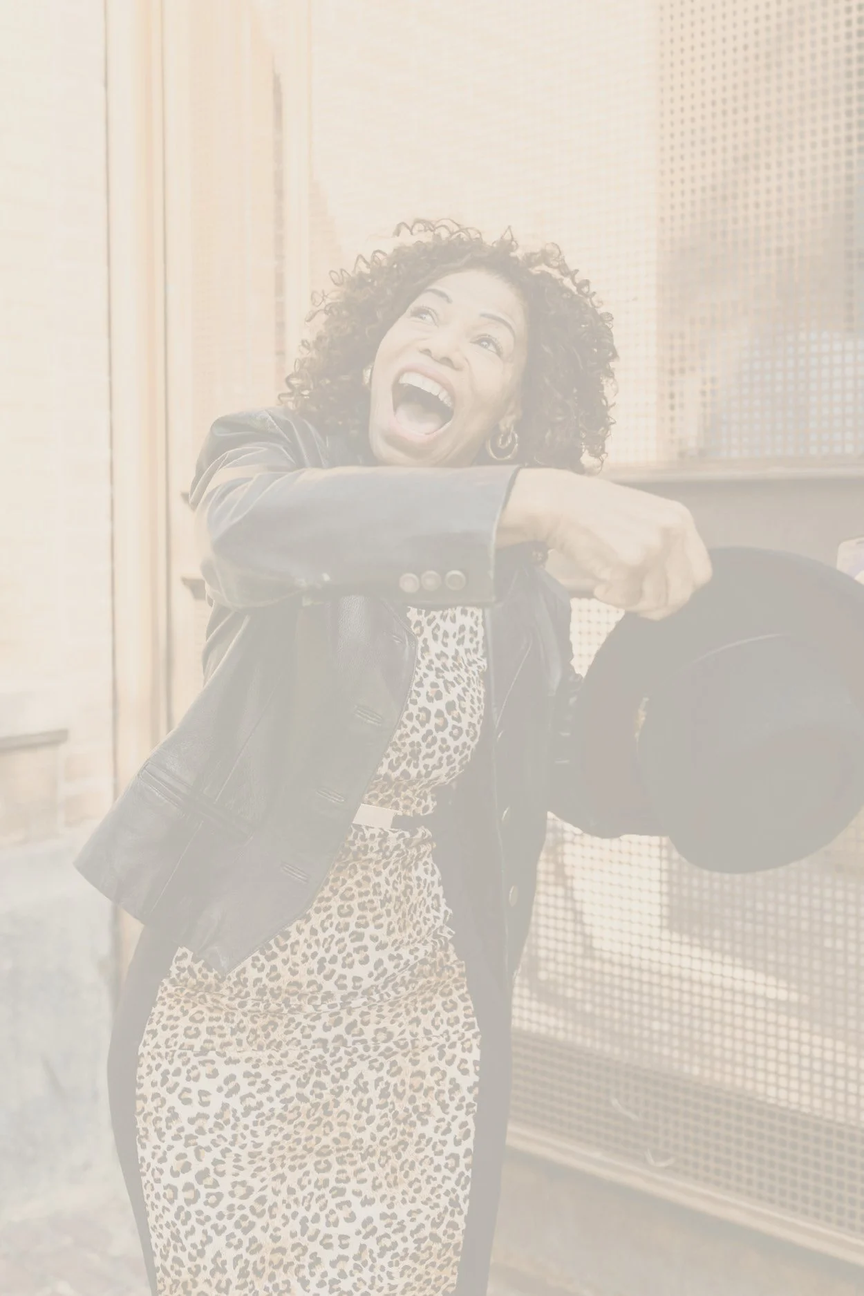 A woman in a leopard print dress and black leather jacket holding a bowler hat, smiling with excitement, standing outside near a building.