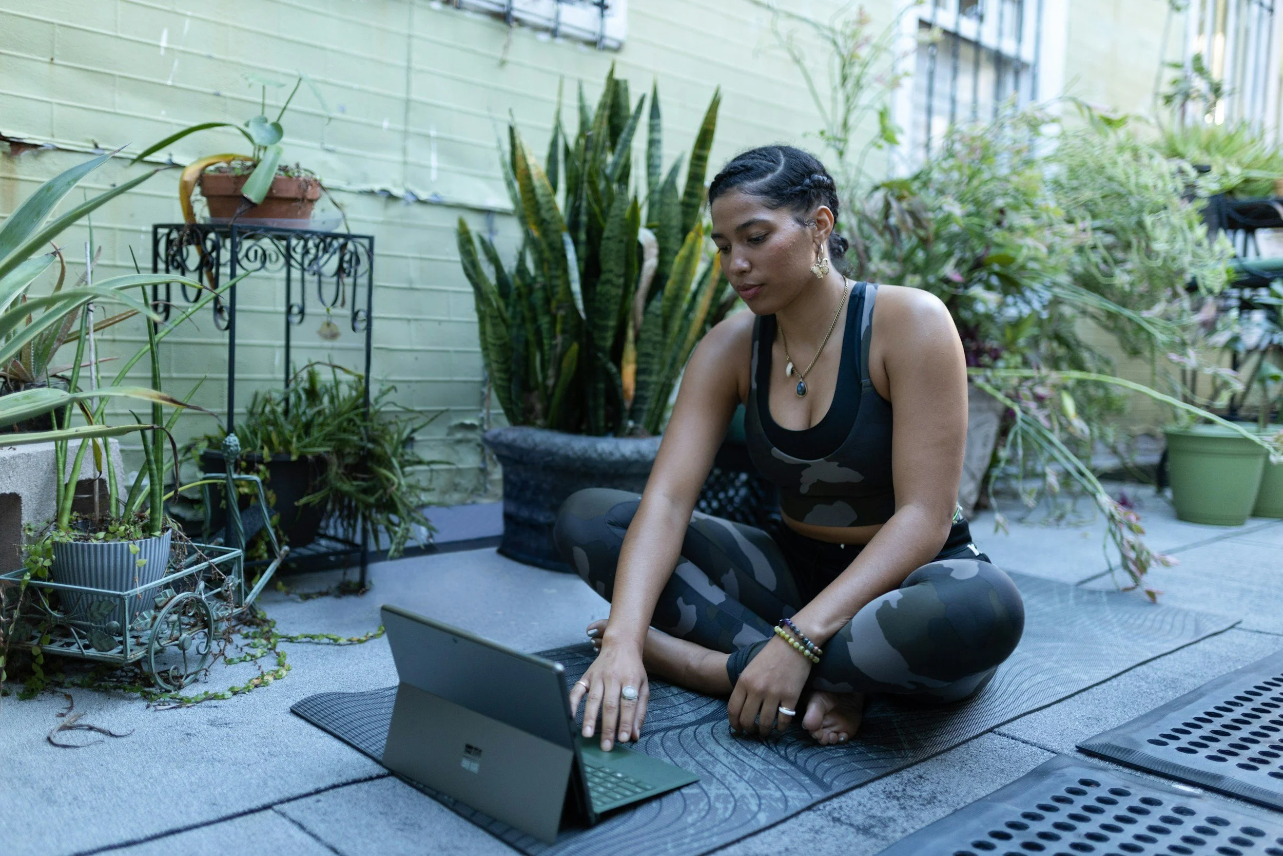 A woman sitting cross-legged on a yoga mat outside, using a Microsoft Surface tablet. She is surrounded by potted plants against a green painted brick wall, wearing black athletic wear with camouflage pattern, jewelry, and has braided hair.