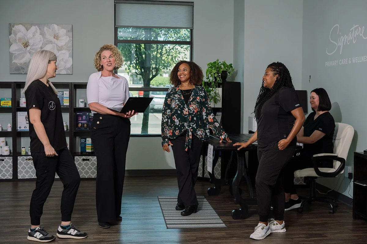 Five women in a professional office setting engaged in conversation, with shelves of medication bottles and a large window in the background.