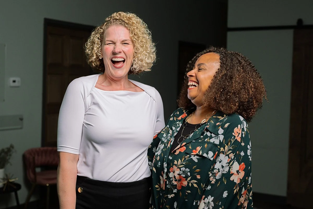 Two women sharing a laugh indoors, one with curly blonde hair wearing a white top, and the other with curly dark hair wearing a floral blazer.