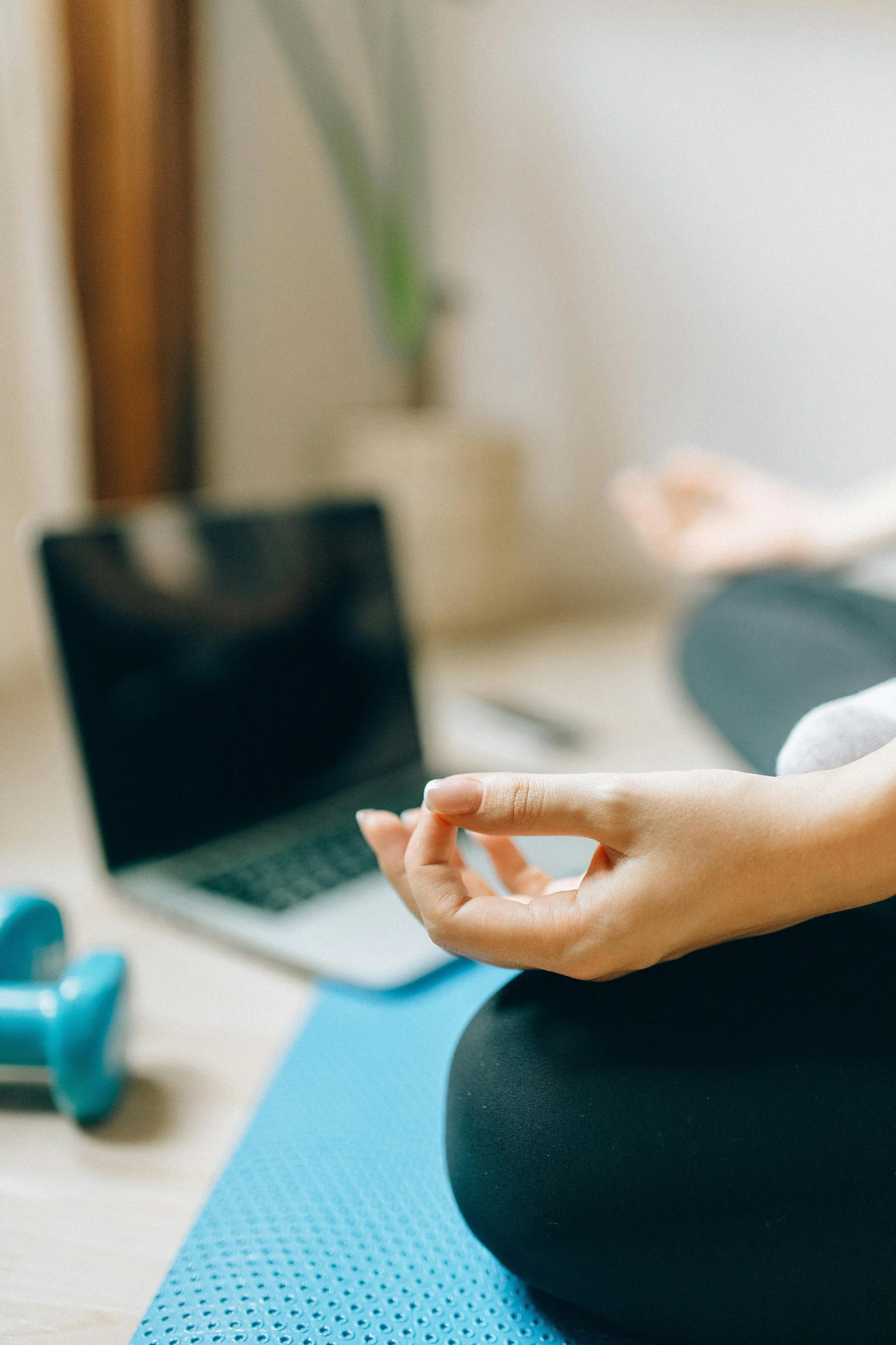 A person sitting on a blue yoga mat practicing meditation with a laptop and small blue dumbbells nearby in a well-lit indoor space.