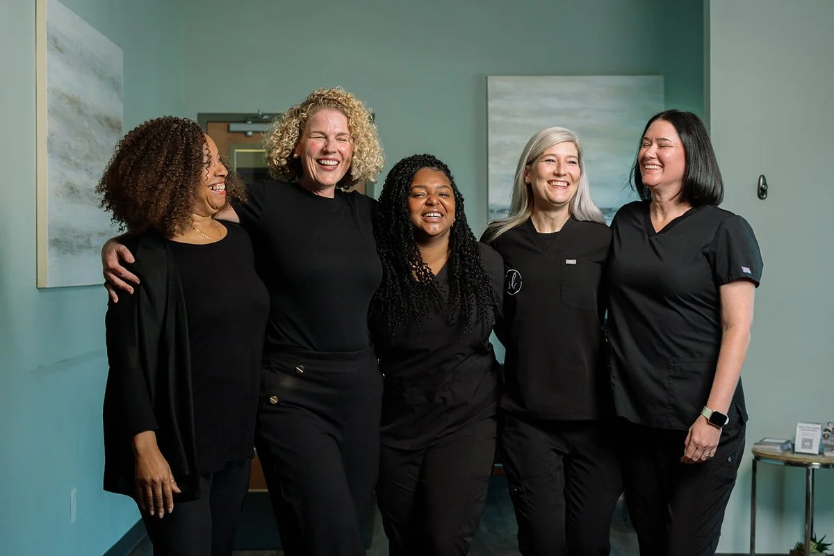 Five women standing together indoors, smiling and laughing, wearing black professional attire, with light green walls and abstract artwork in the background.