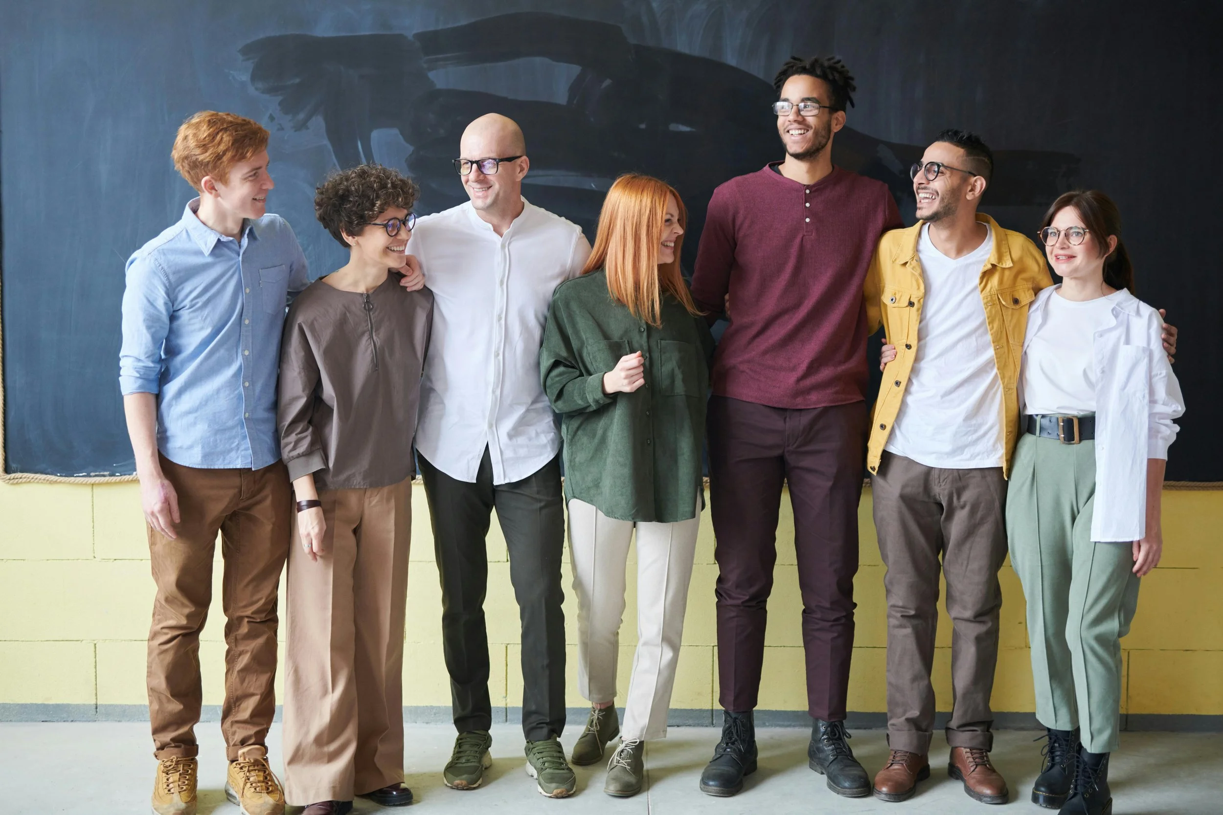 Group of seven diverse young adults standing together in front of a chalkboard, smiling and talking in a classroom or academic setting.