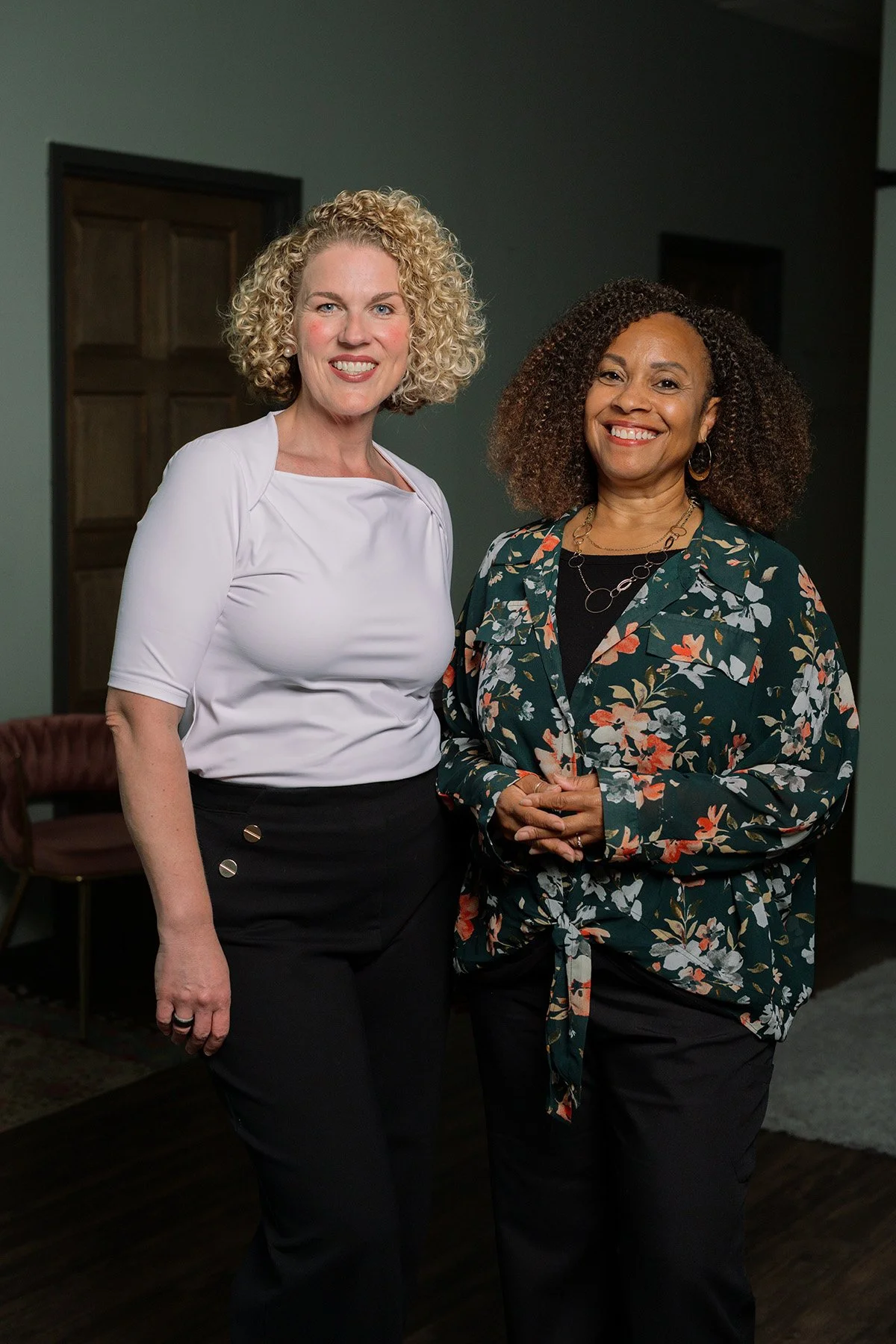 Two women standing indoors, smiling at the camera, with a wooden door and green wall in the background.