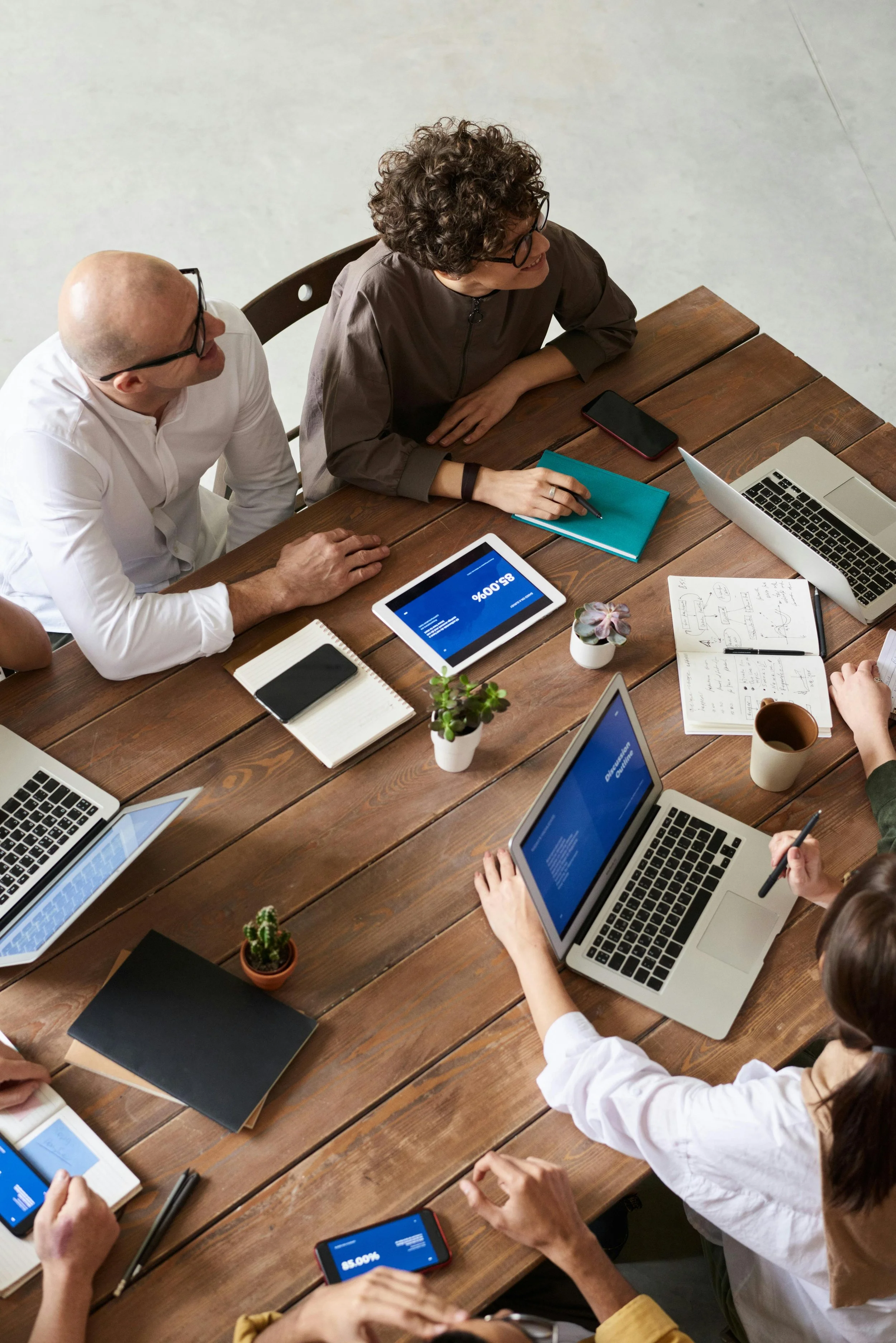 A group of people having a business meeting around a wooden table with laptops, tablets, notebooks, and cups.