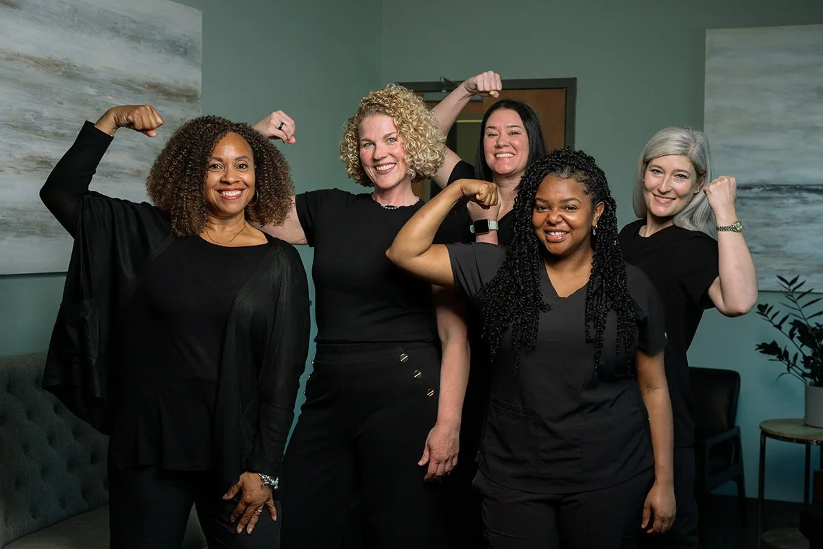 Five women standing together in a room, flexing their biceps and smiling confidently, dressed in black, showing strength and empowerment.