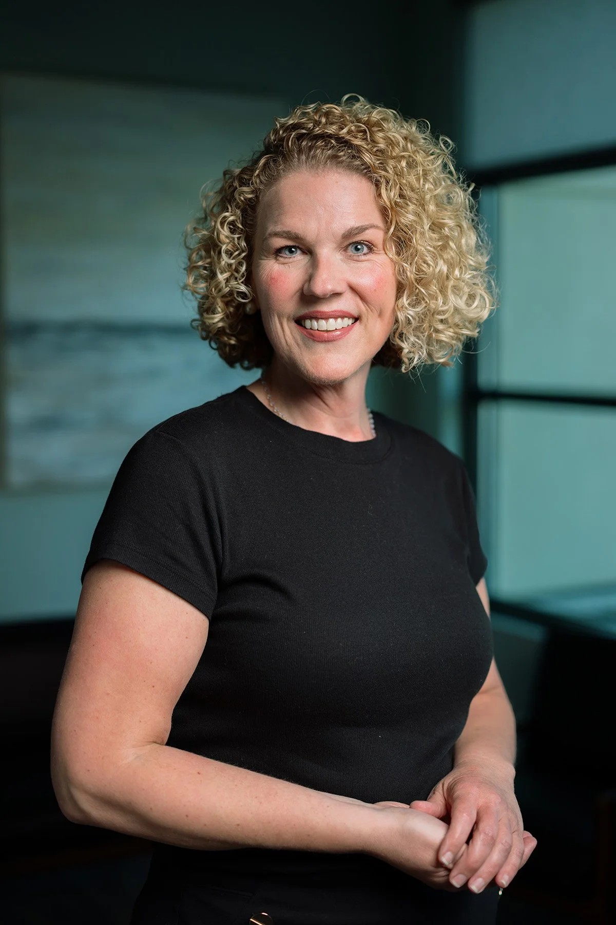 A woman with curly blonde hair wearing a black shirt, smiling in an indoor setting with a window in the background.