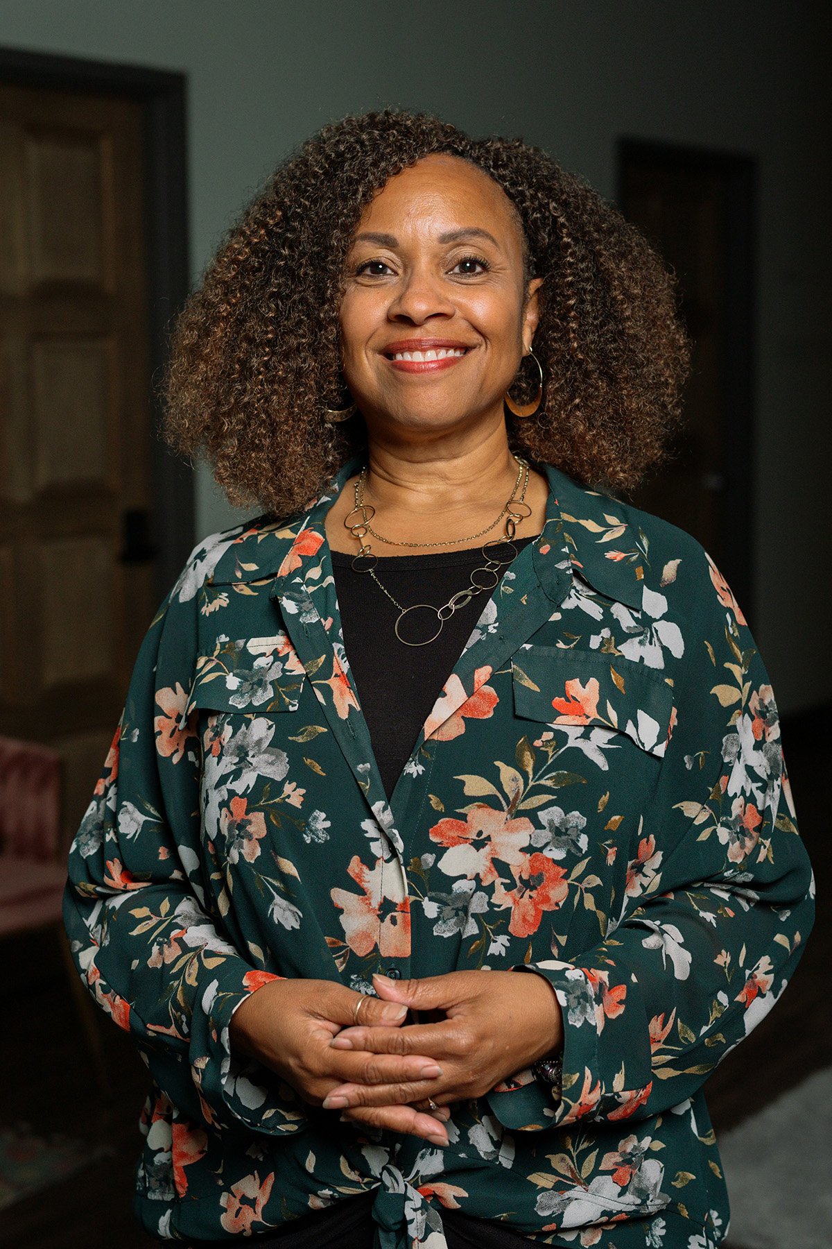 A woman with curly hair in a floral blouse and jewelry standing in a room.