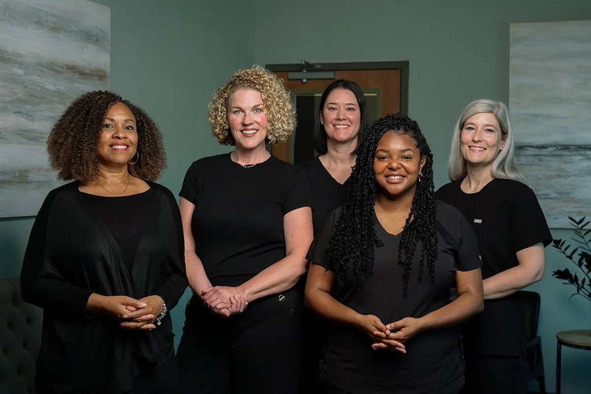Five women standing together indoors, smiling, with abstract art paintings on the wall behind them.