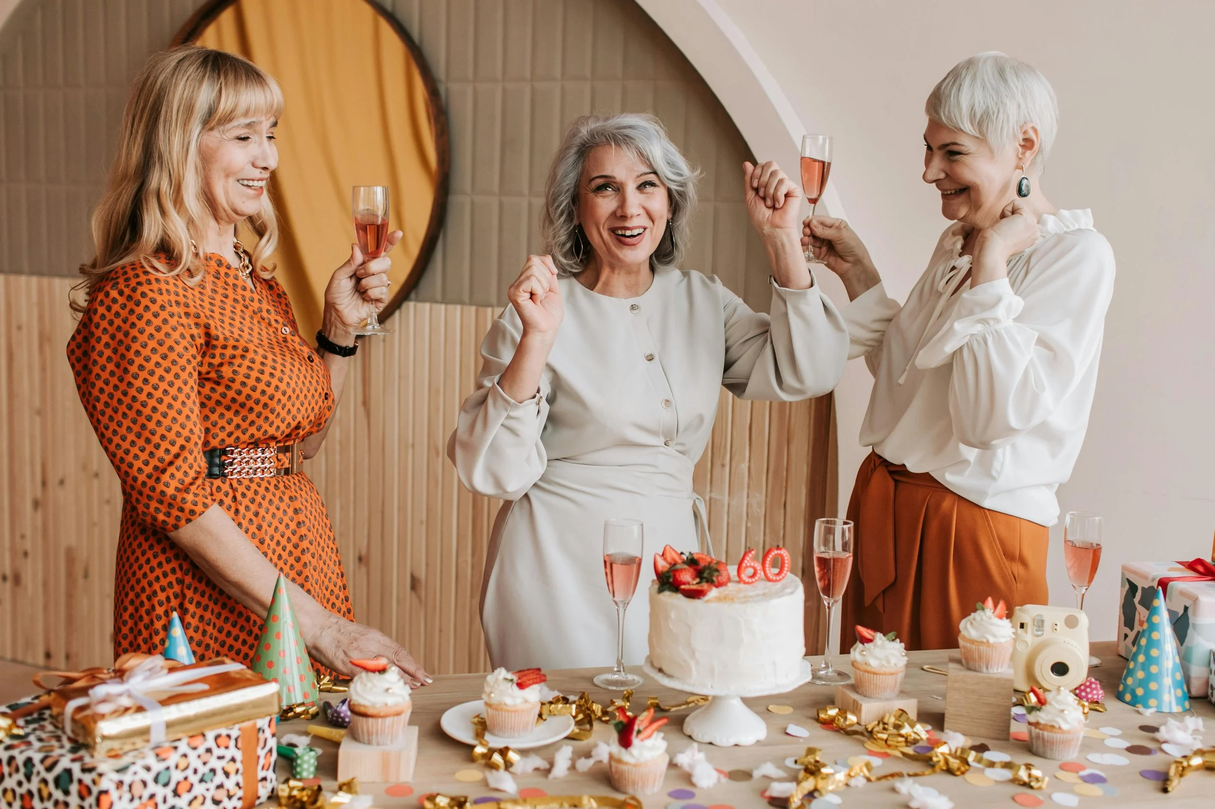 Women celebrating a 60th birthday with a decorated cake, cupcakes, confetti, and party hats, holding glasses of pink sparkling beverage.