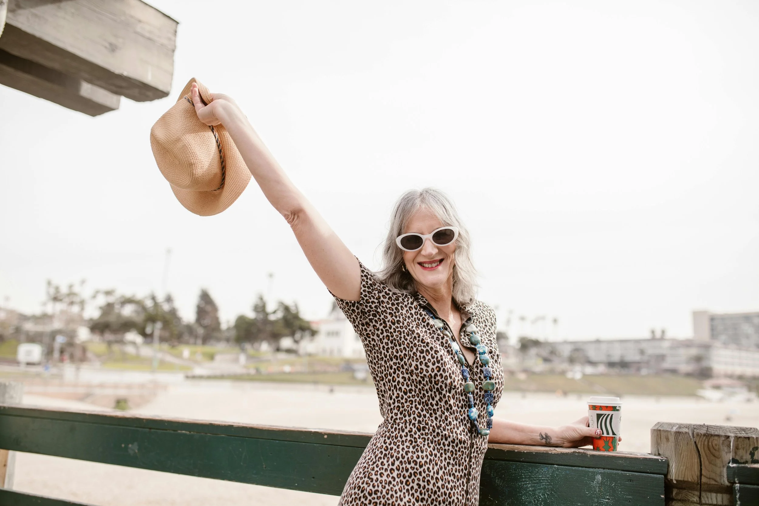 Smiling elderly woman wearing sunglasses and a leopard print dress, holding a straw hat in one hand and a coffee cup in the other, standing outdoors at a park or beach walkway.
