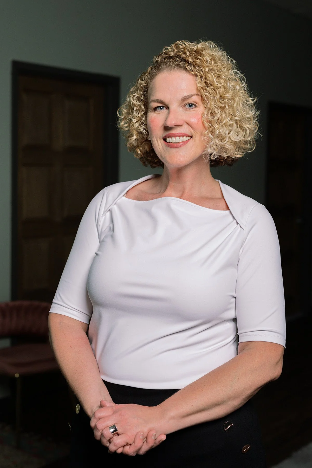 A woman with shoulder-length curly blonde hair wearing a white top smiling and standing indoors with a dark background.