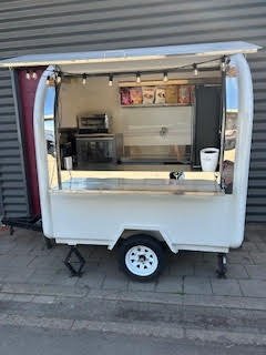 Trailer de food truck blanc avec une fenêtre ouverte, intérieur visible avec une caisse et une petite étagère, stationné sur le trottoir.