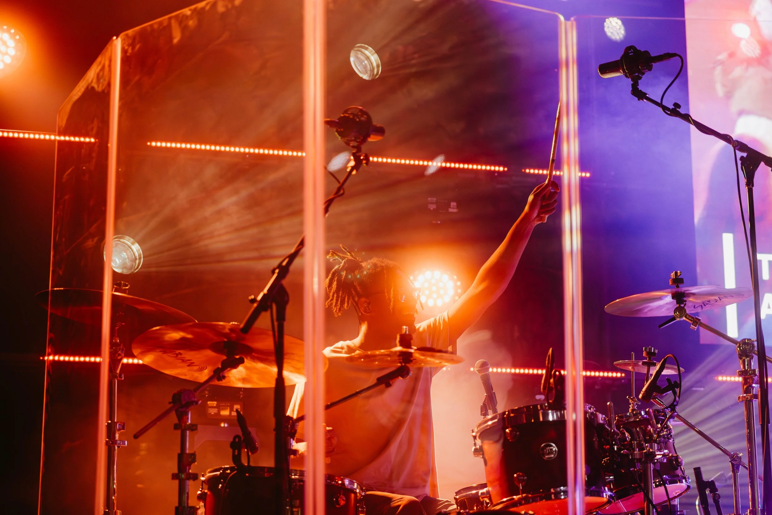 A drummer playing behind a transparent shield on a stage illuminated with orange and purple lights, with microphone stands nearby.