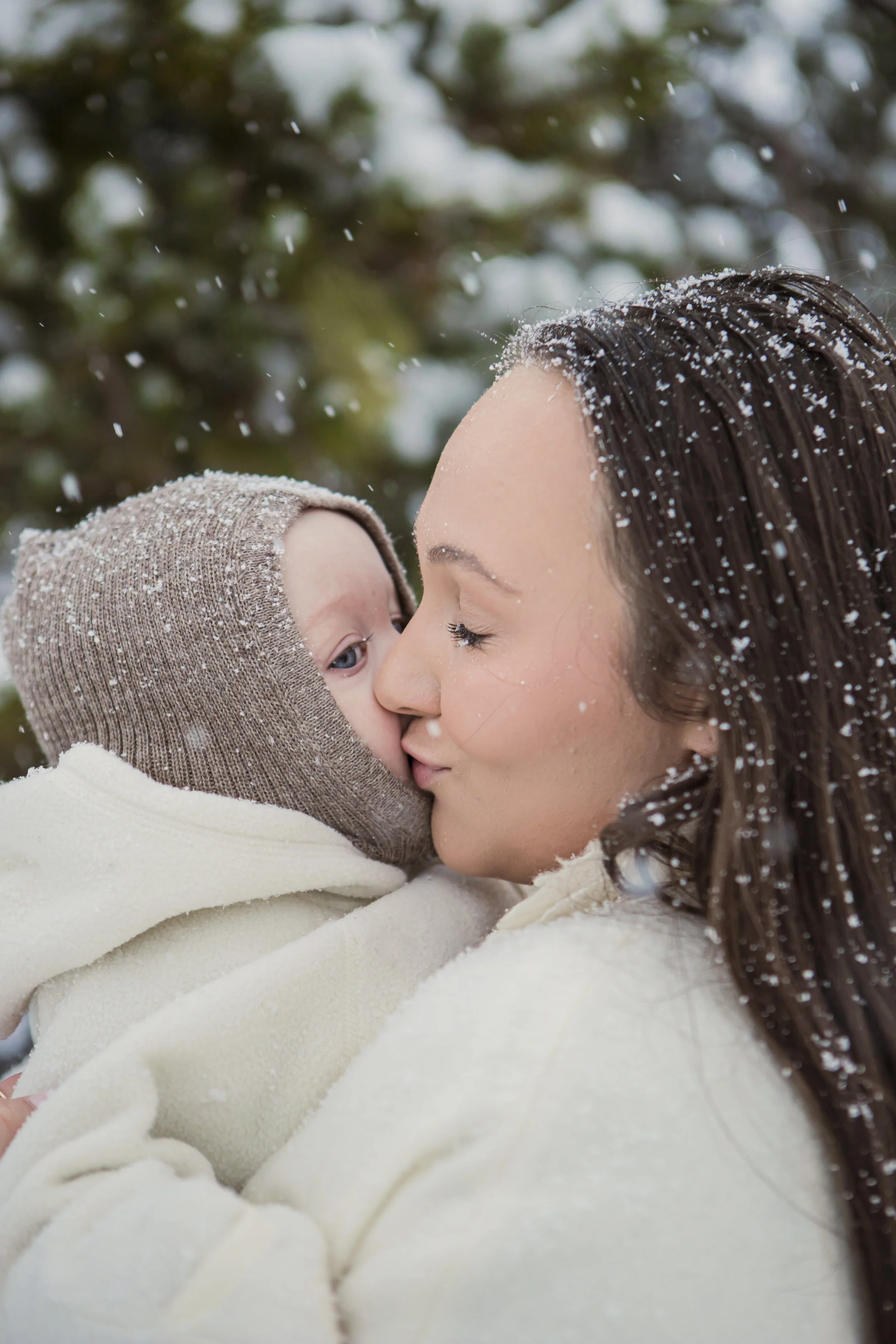 A woman holding a child close, both with snowy weather, near snow-covered trees, sharing a tender moment.