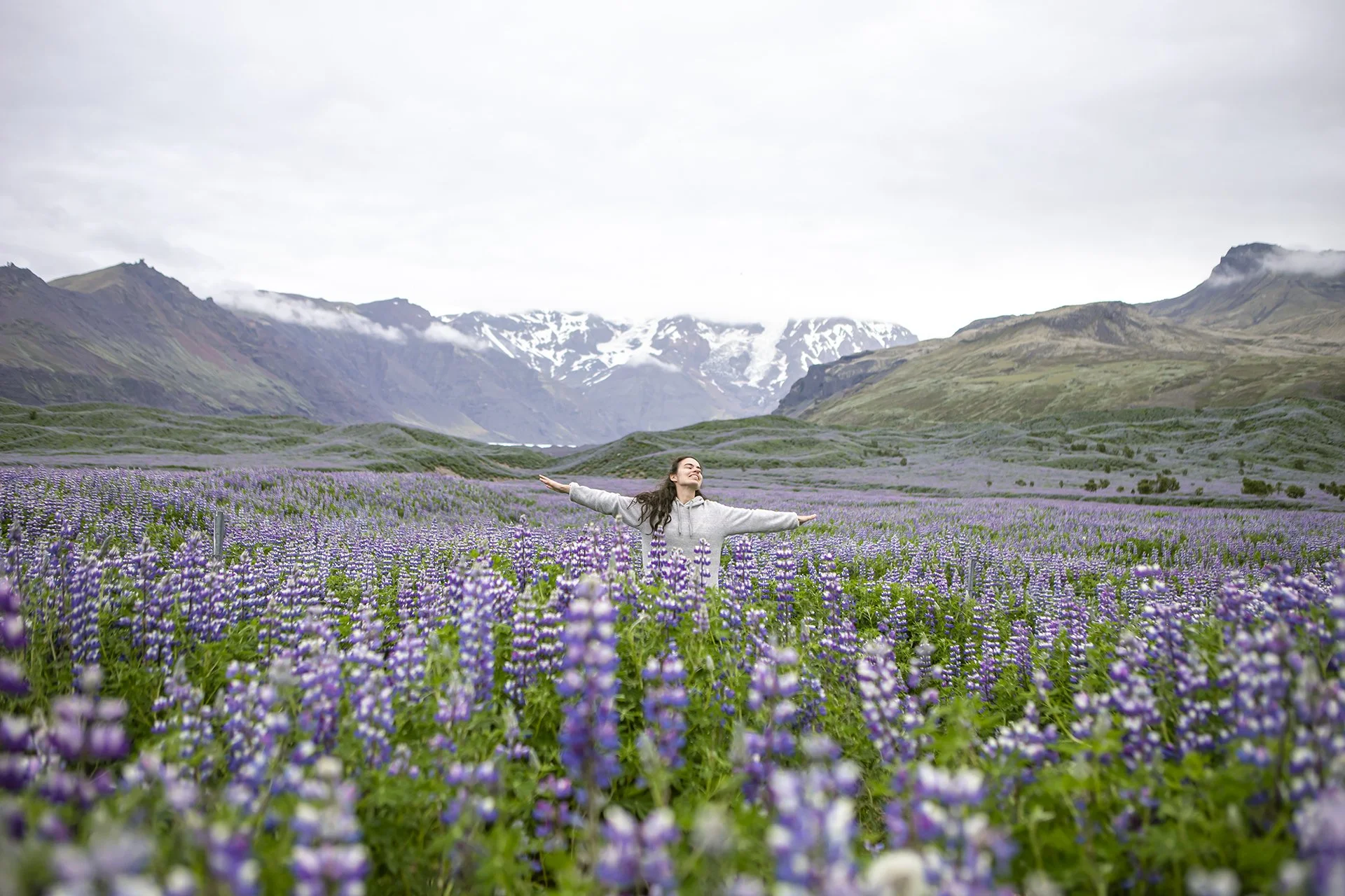 A woman with outstretched arms standing in a vast field of purple wildflowers, with mountain ranges in the background under a cloudy sky.