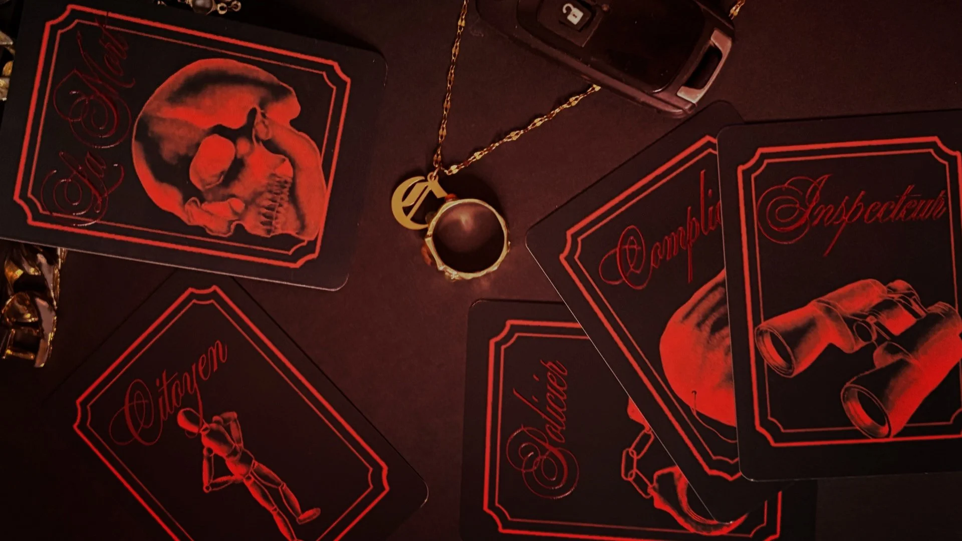 Des cartes de jeu en noir et rouge avec des illustrations et des textes comme "Citoyen", "Inspecteur", et "La Mort", un bijou avec une chaîne et une clé de voiture sur une surface sombre.