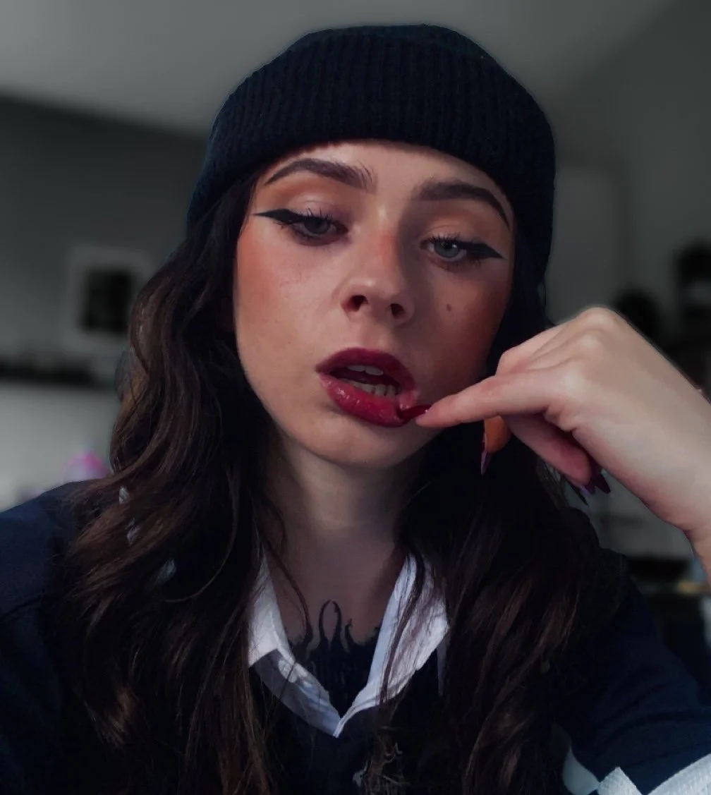 Femme portant un bonnet noir et une chemise blanche avec un pull, se tenant le menton, maquillage d'yeux prononcé et rouge à lèvres rouge foncé.
