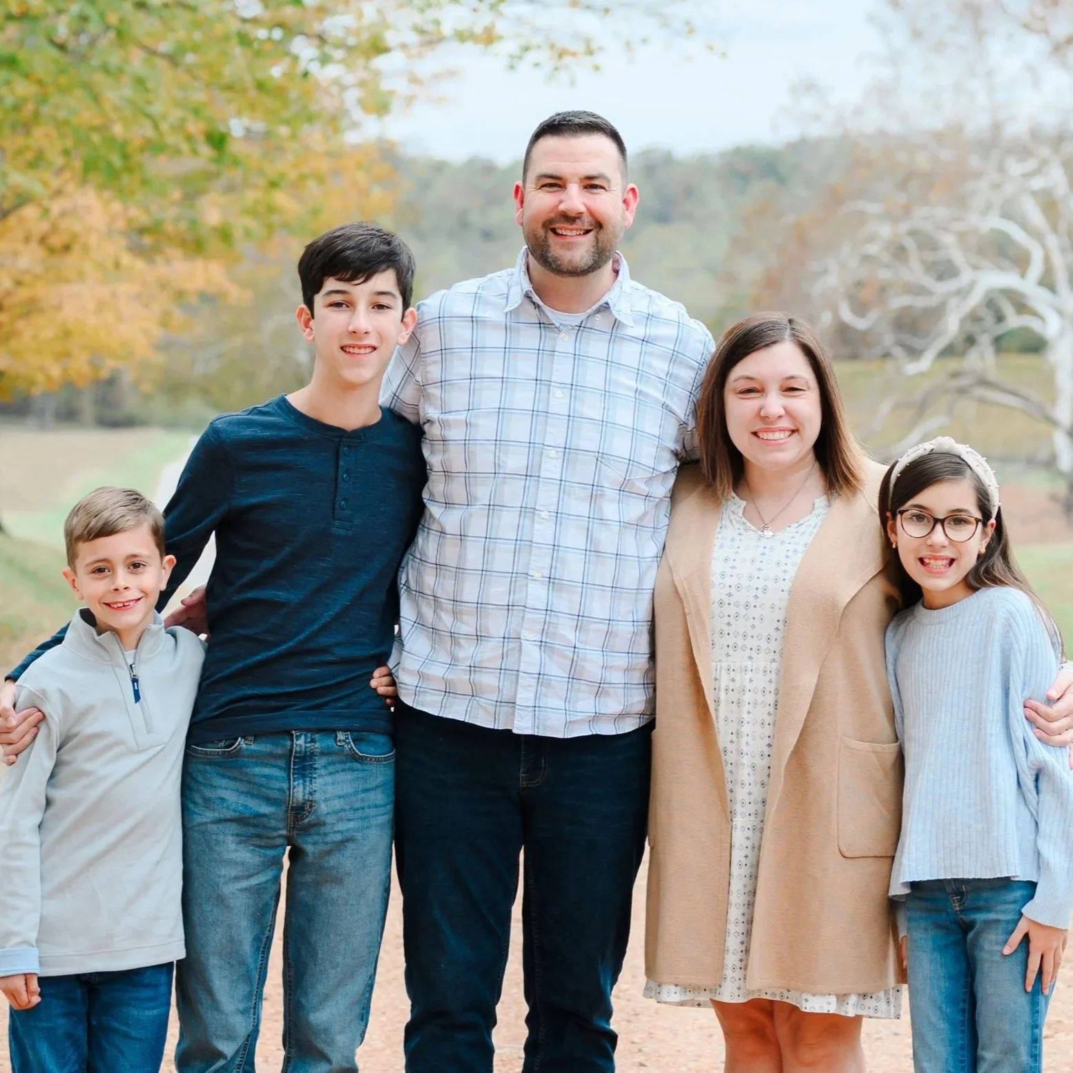 A family of six posing outdoors in a wooded area with trees and fallen leaves on the ground. The family members include two adults, two boys, and two girls, all smiling and dressed casually.