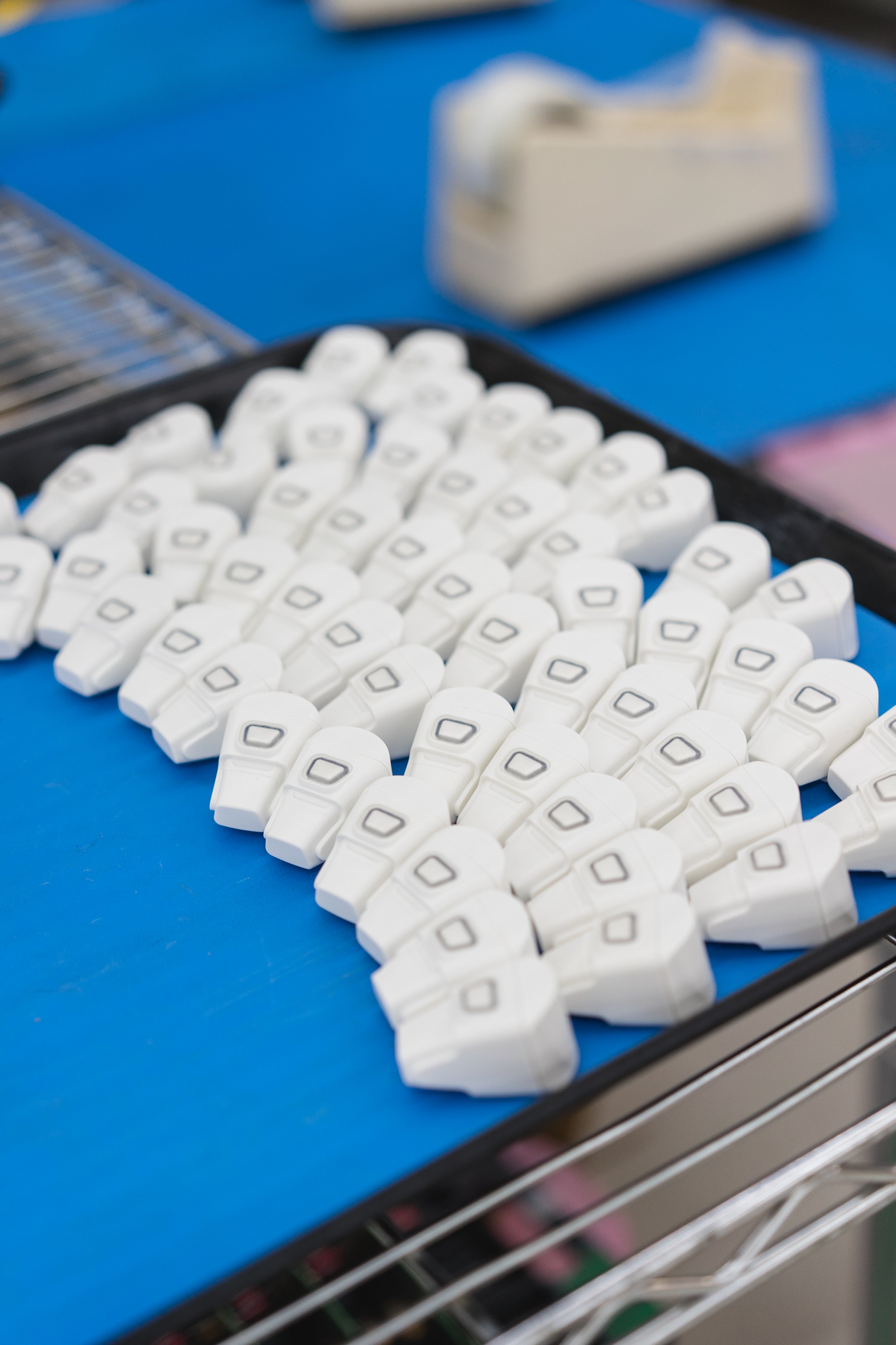 A tray of white devices on a blue surface, with a blurry background of a tape dispenser and a wire rack.