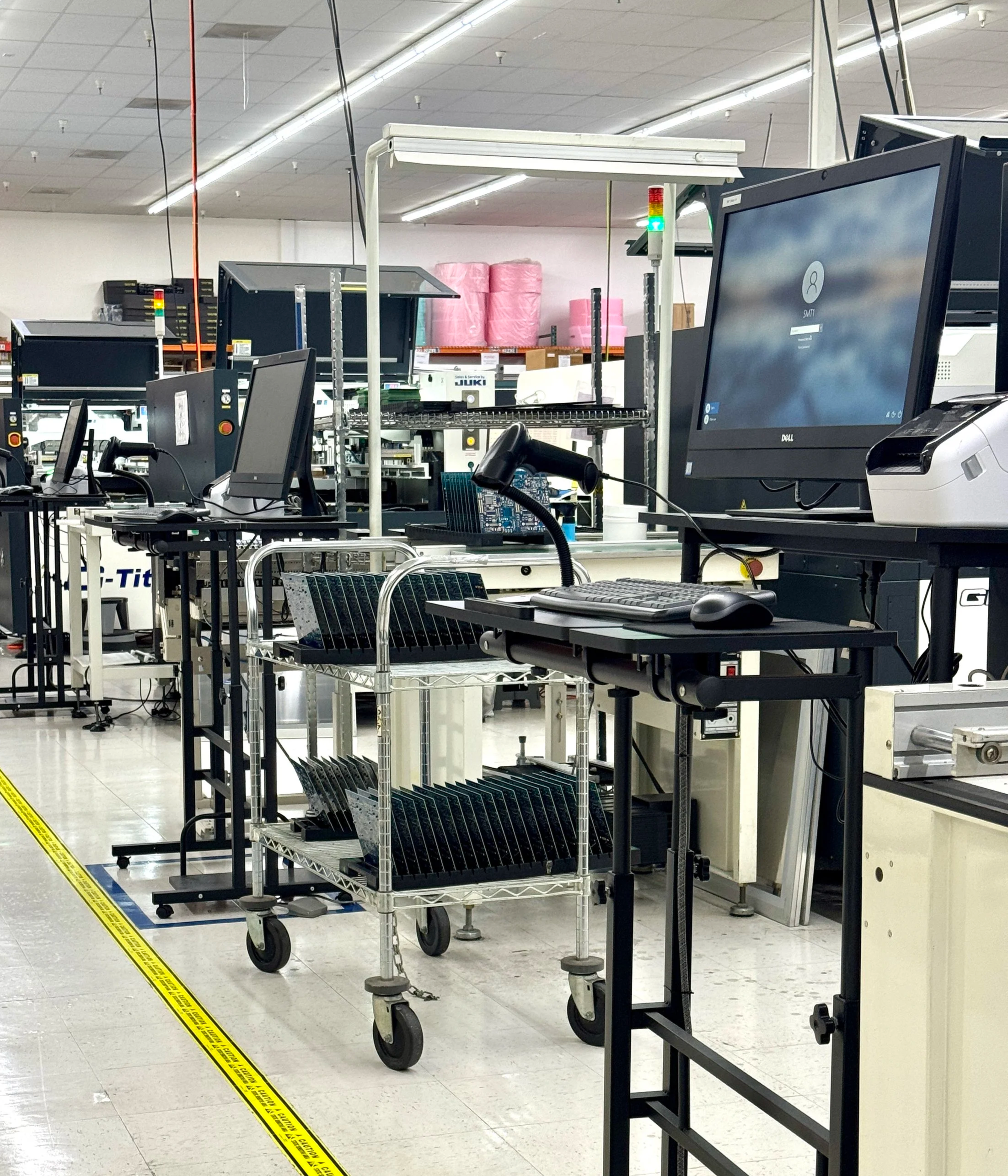 Scanning stations with computer monitors, keyboards, and other electronics on black counters in a large manufacturing facility, with a yellow caution tape on the floor and warehouse shelves with pink packing materials in the background.