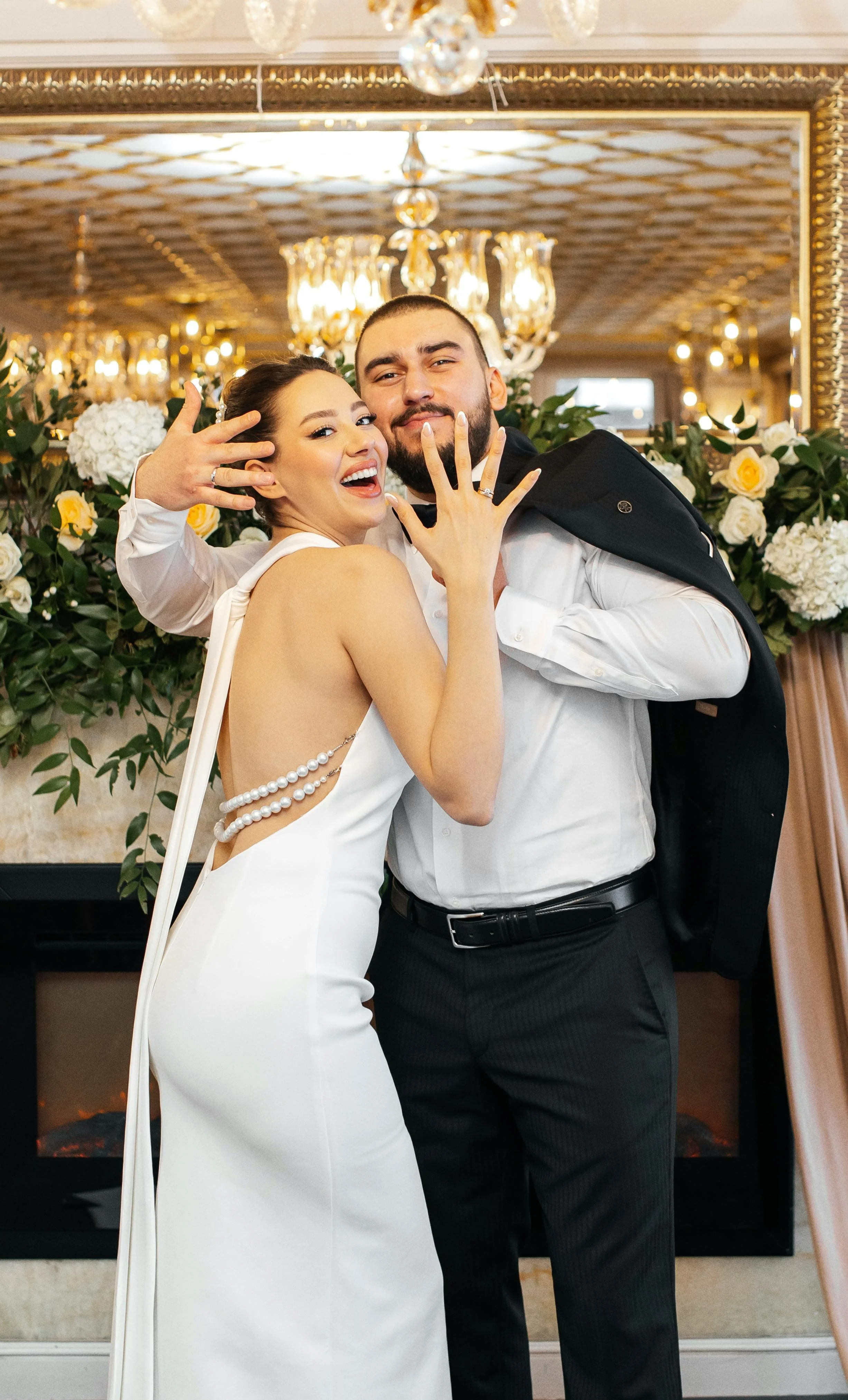 A bride and groom smiling and showing off their wedding bands at their wedding reception in front of a decorated mantel.
