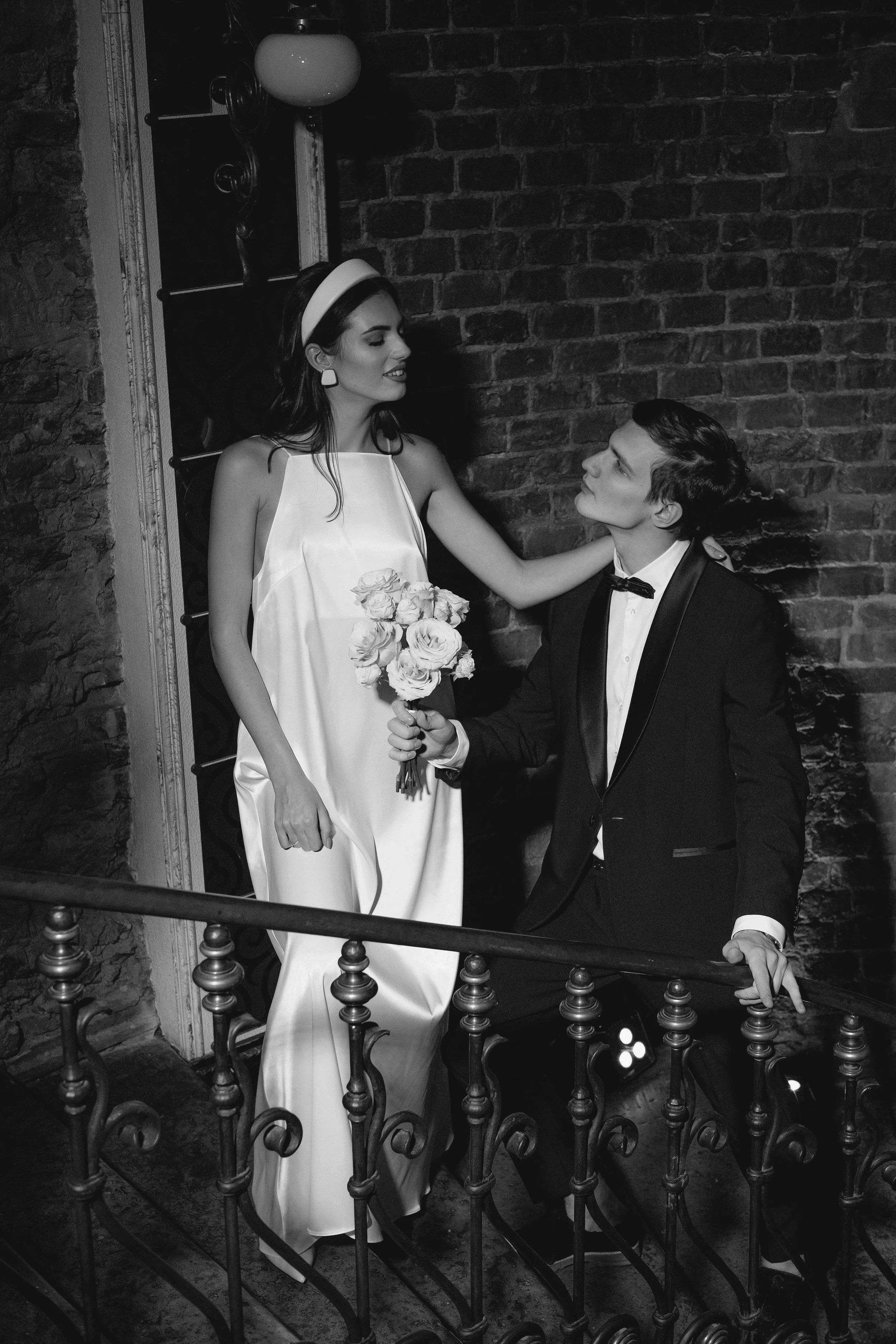 Bride and groom on a balcony together, exchanging soft looks and a bouquet of roses.