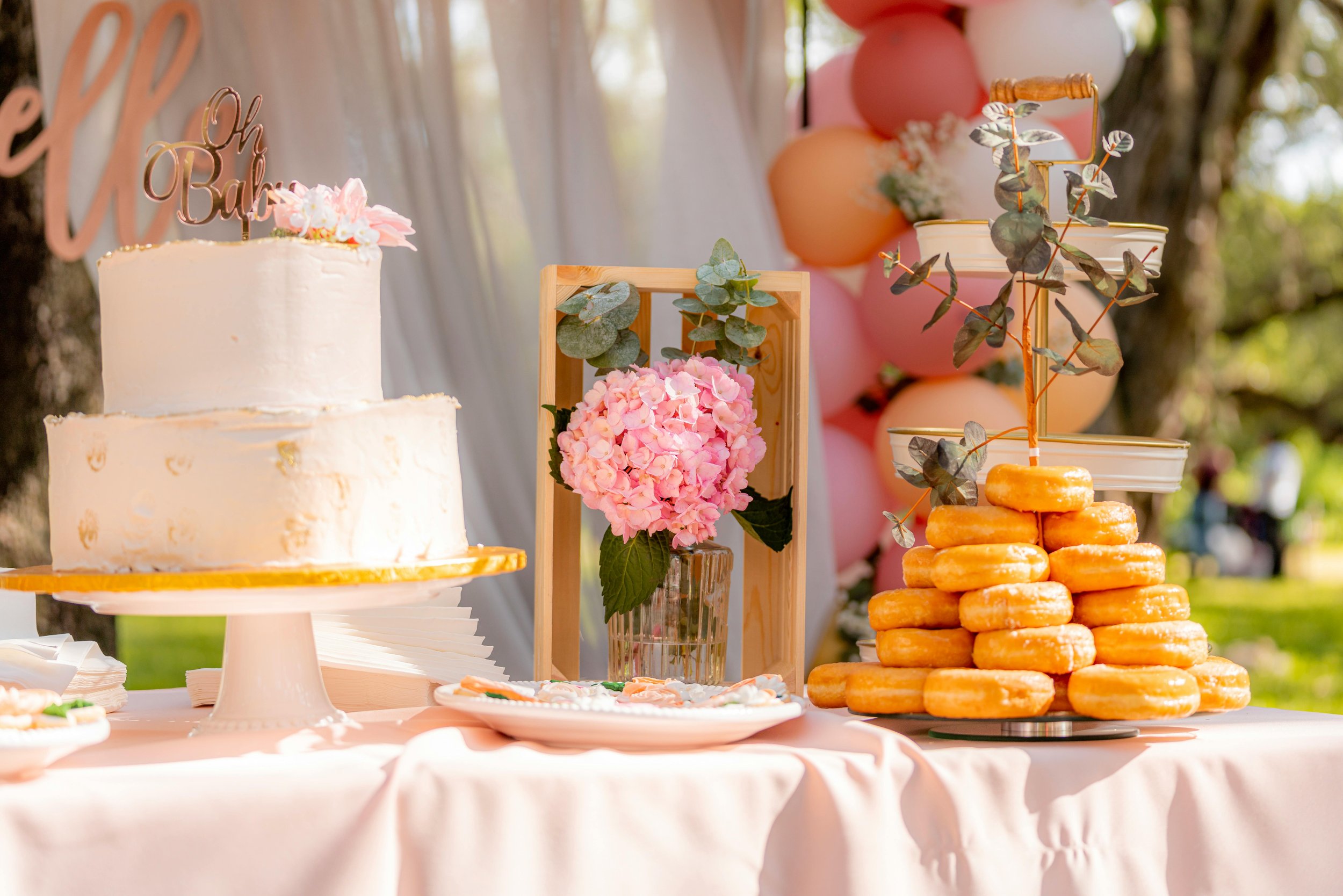 A dessert table dressed in a pink tablecloth with a white two-tier cake, pink florals, and a donut stand.