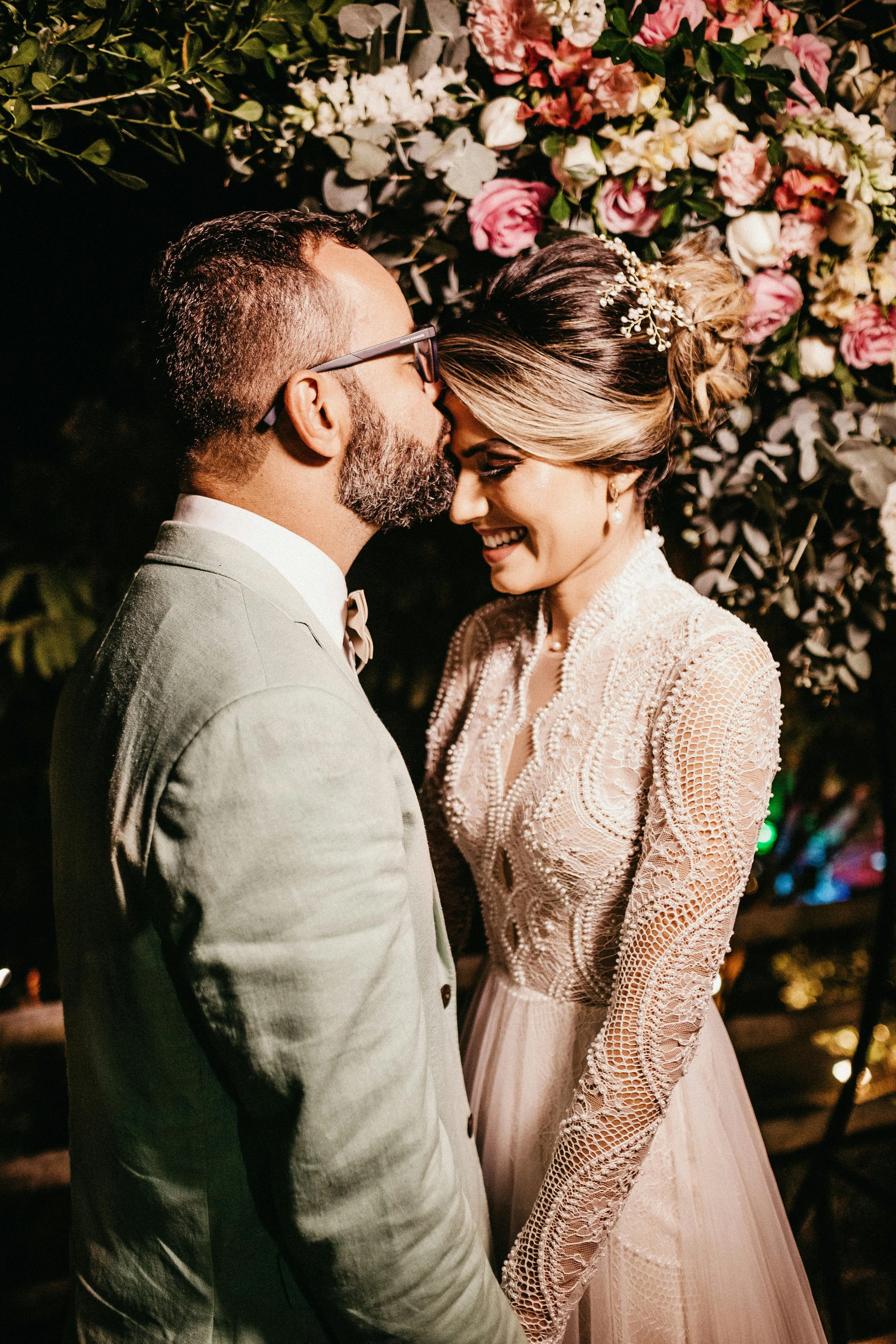 A bride dressed in pink surrounded by pink florals getting kissed on the forehead as she smiles.