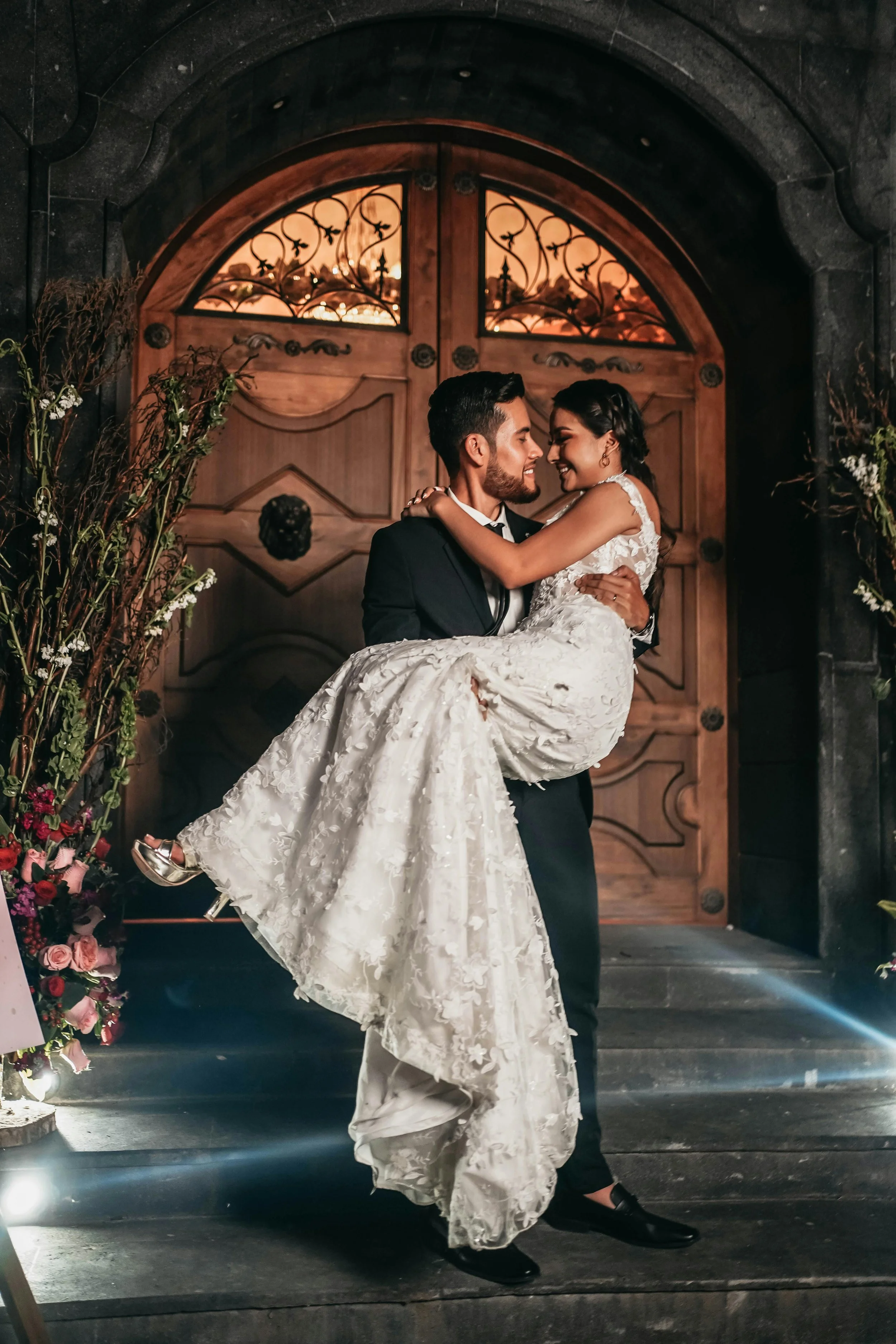 A groom is carrying his bride outside a venue with big brown entrance doors.
