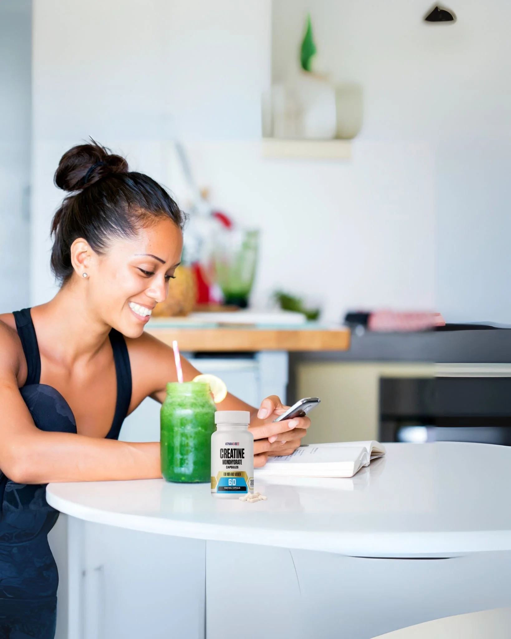 A woman with dark hair in a bun smiling while looking at her phone, sitting at a white table with a green smoothie and a bottle of creatine supplement.