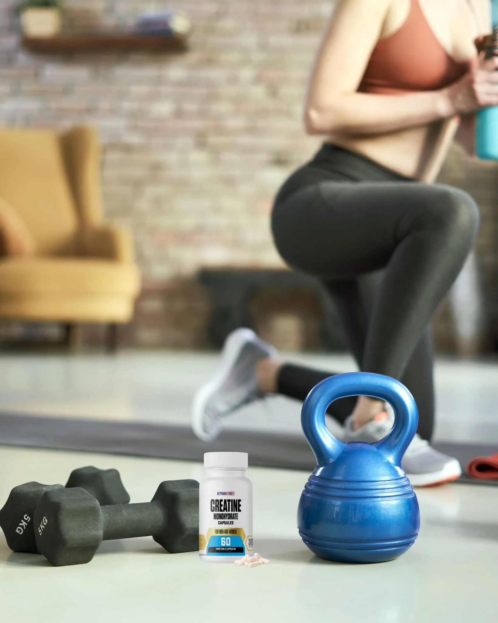 Dumbbells, creatine supplement capsule bottle, and a blue kettlebell on the floor with a woman stretching in the background.