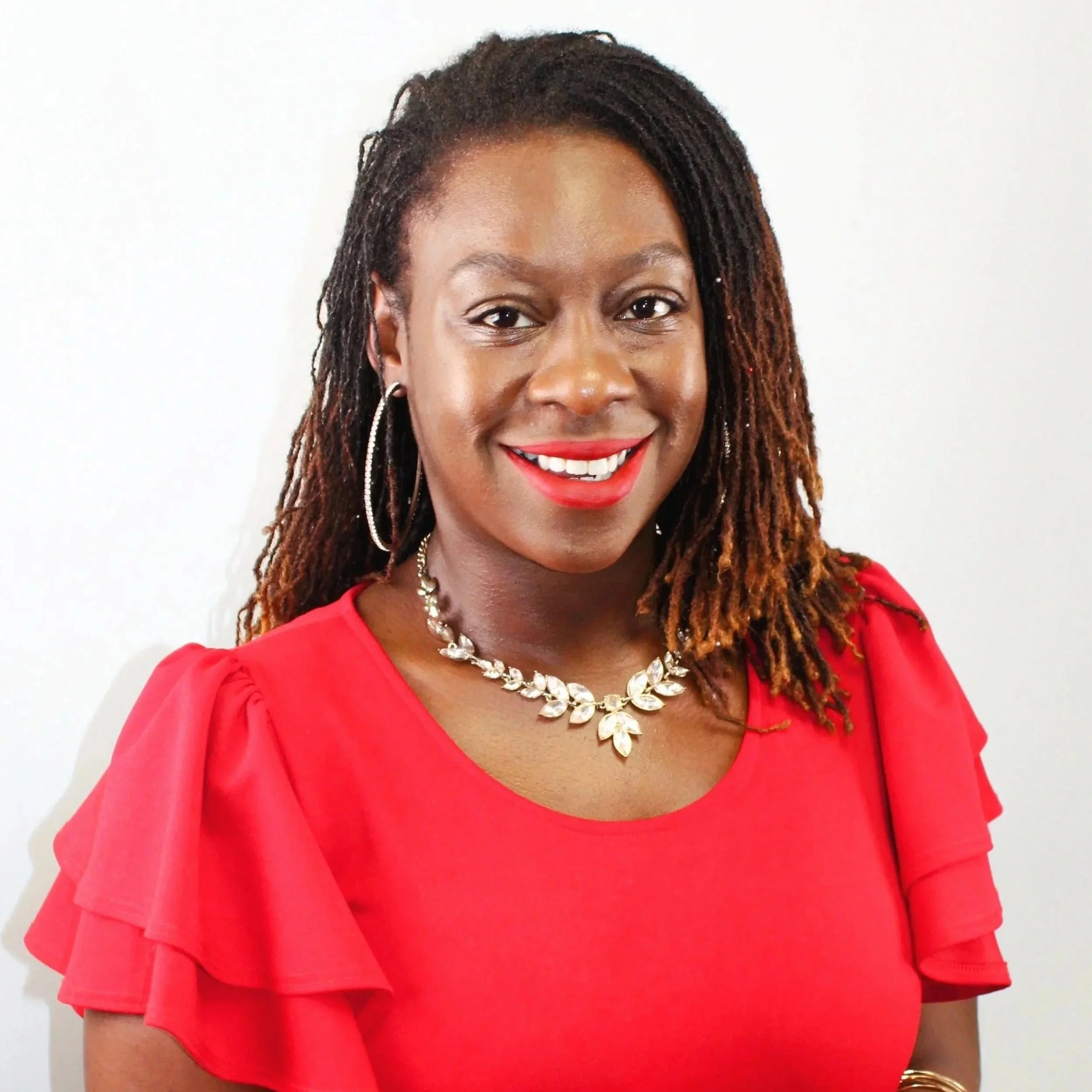 Woman with dark hair and brown skin, wearing a red dress with ruffled sleeves, a silver statement necklace, hoop earrings, and red lipstick, smiling against a plain white background.